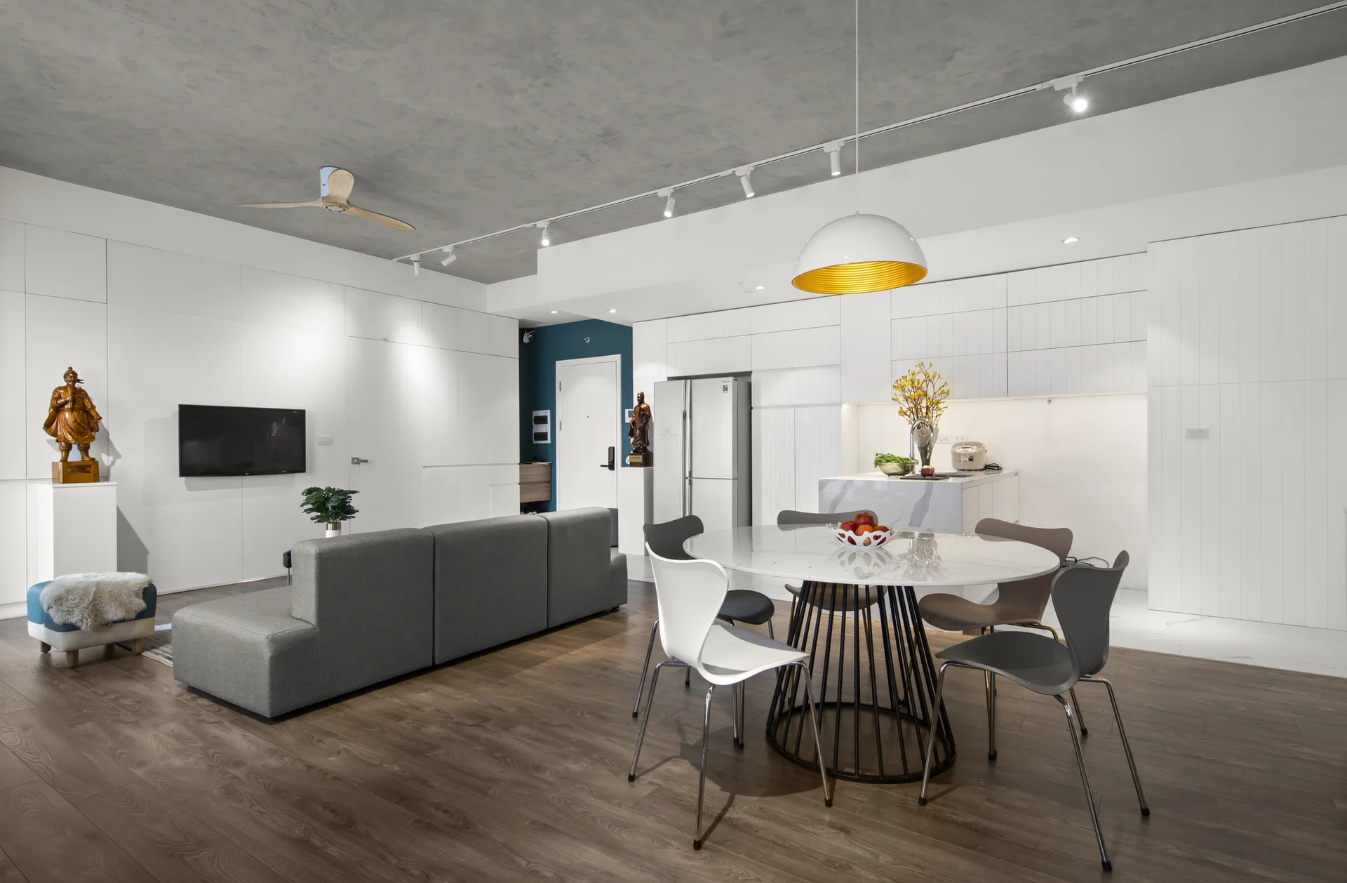 An open-plan living and dining space featuring a grey sofa, a marble dining table, white minimalist cabinetry, and an exposed concrete ceiling.