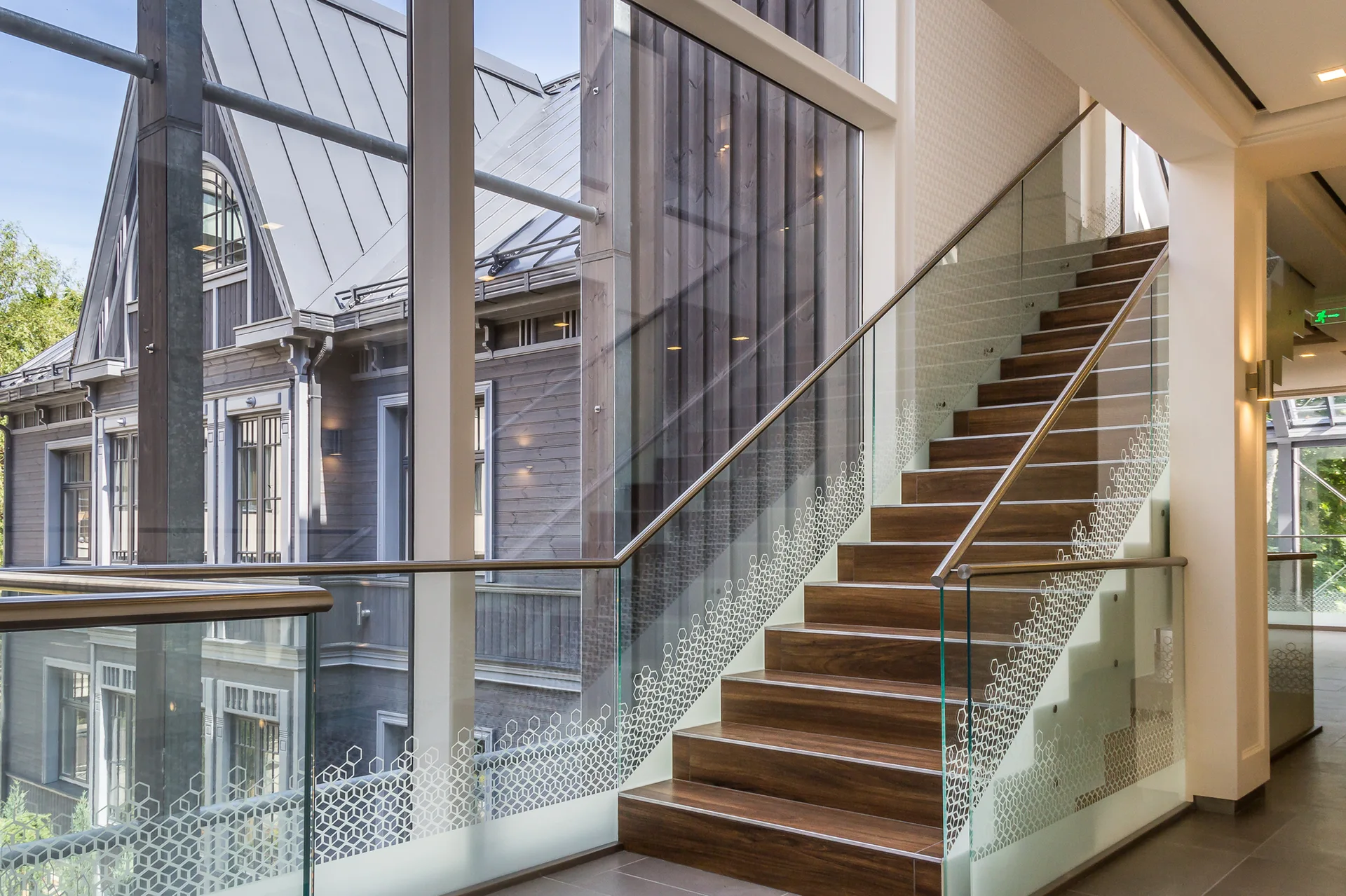 A contemporary wooden staircase with patterned glass railings is situated next to tall windows overlooking an adjacent building.