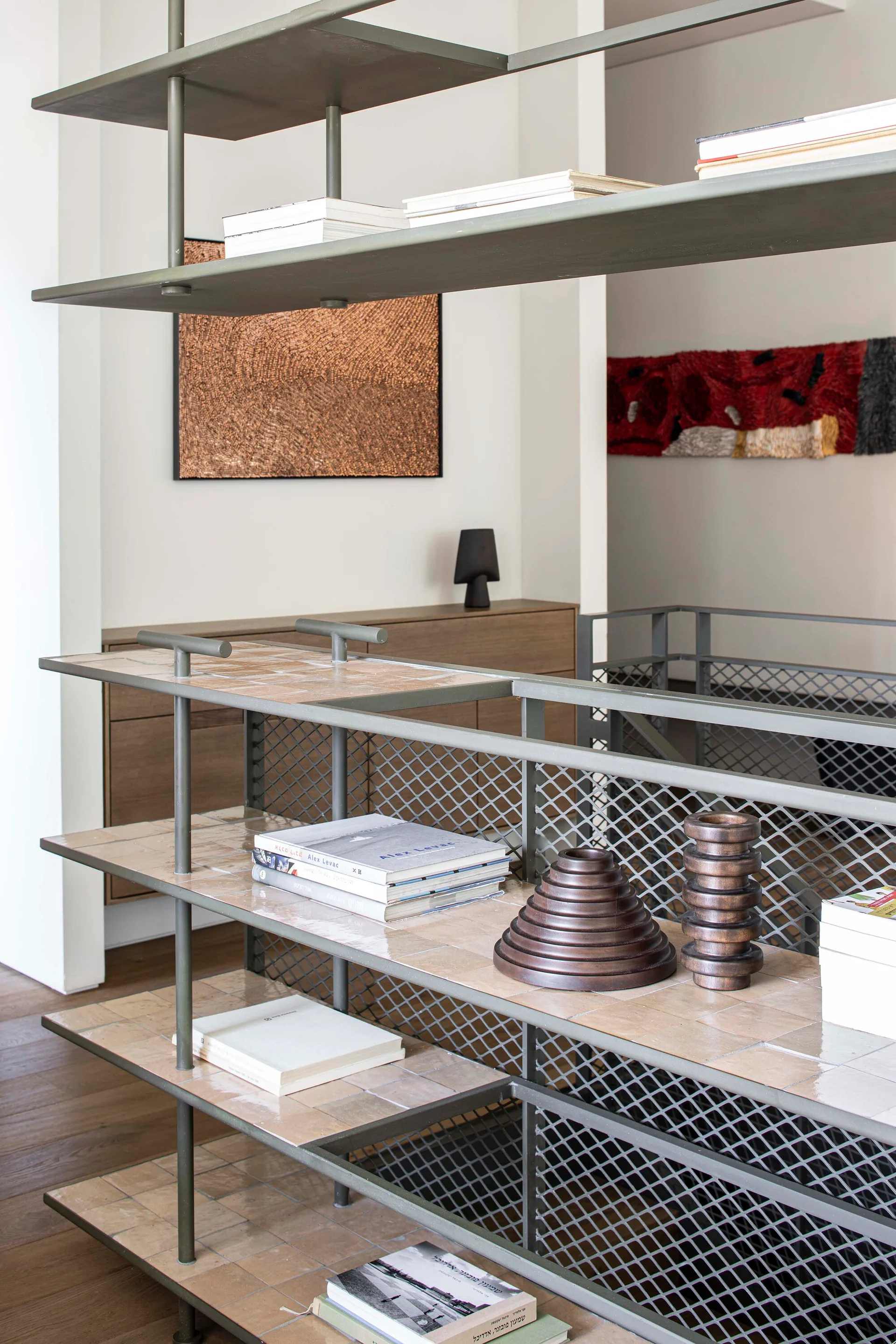 An olive green metal shelving unit features glazed beige tile shelves holding books and wooden geometric sculptures in a modern interior.