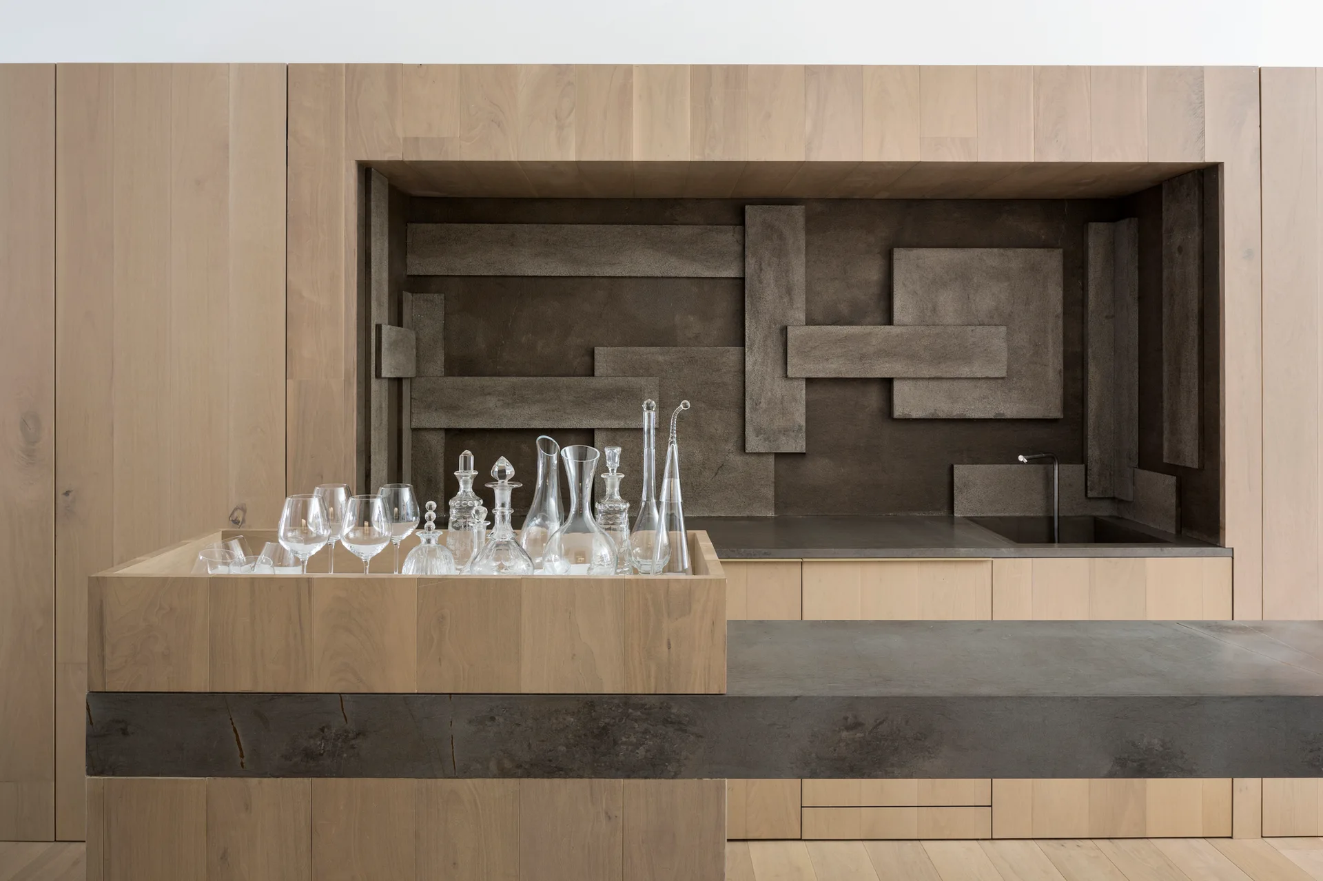 A minimalist kitchen bar features light wood cabinetry, a charcoal countertop, a geometric relief backsplash, and glass decanters in a built-in wooden box.