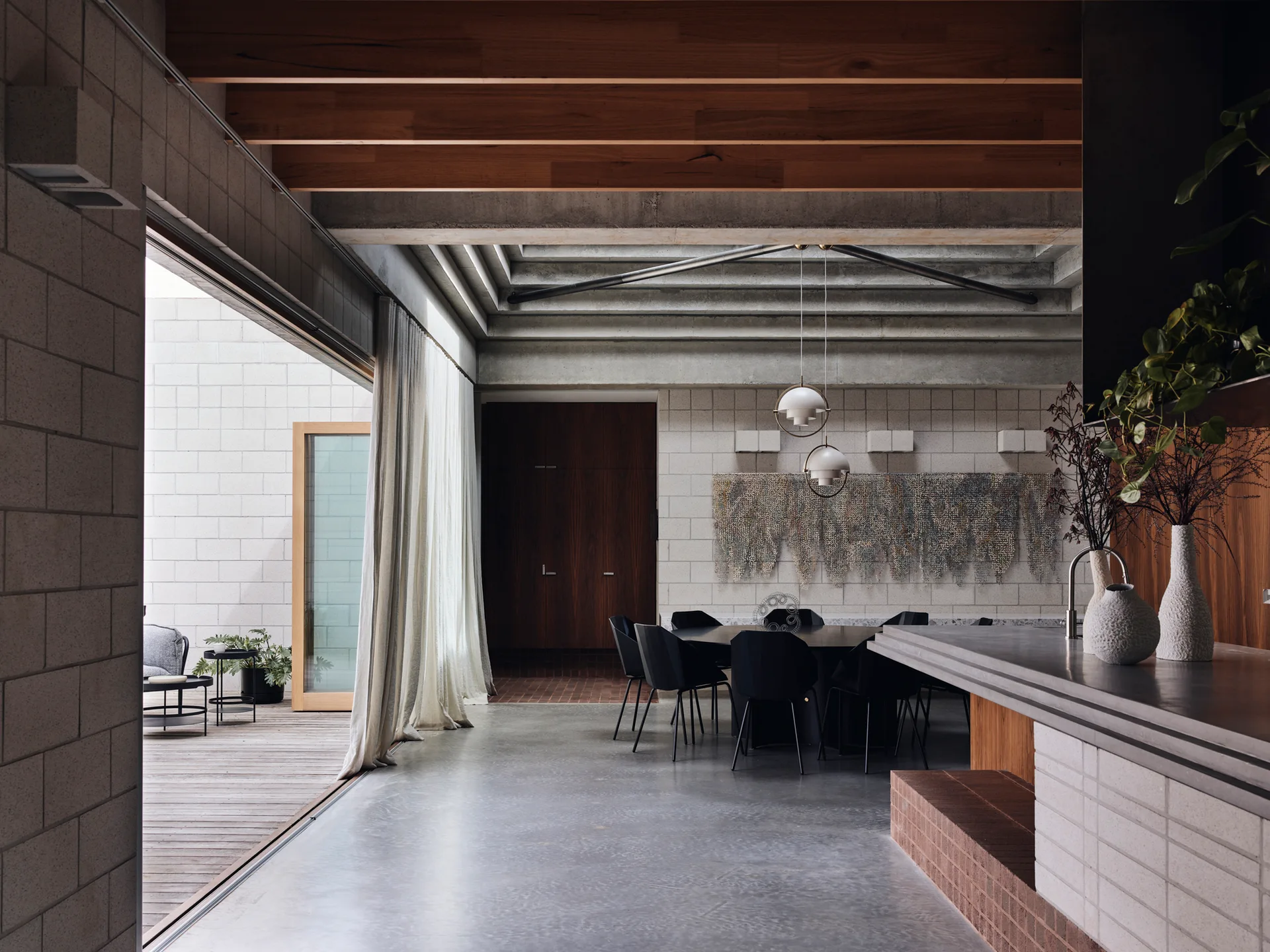 A minimalist dining room featuring a black table and chairs, concrete block walls, timber ceiling beams, and sliding glass doors to a deck.