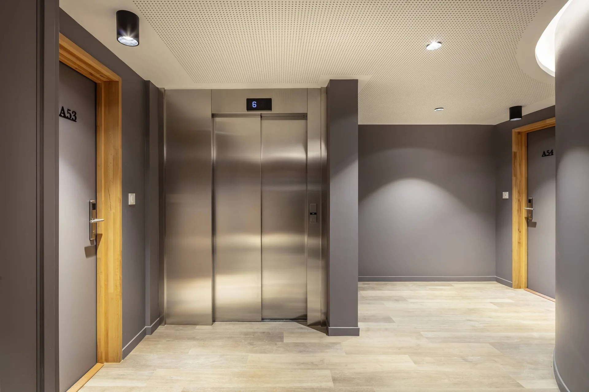 A modern hallway features a central stainless steel elevator, dark gray walls, light wood-framed doors, and light-toned wood plank flooring.