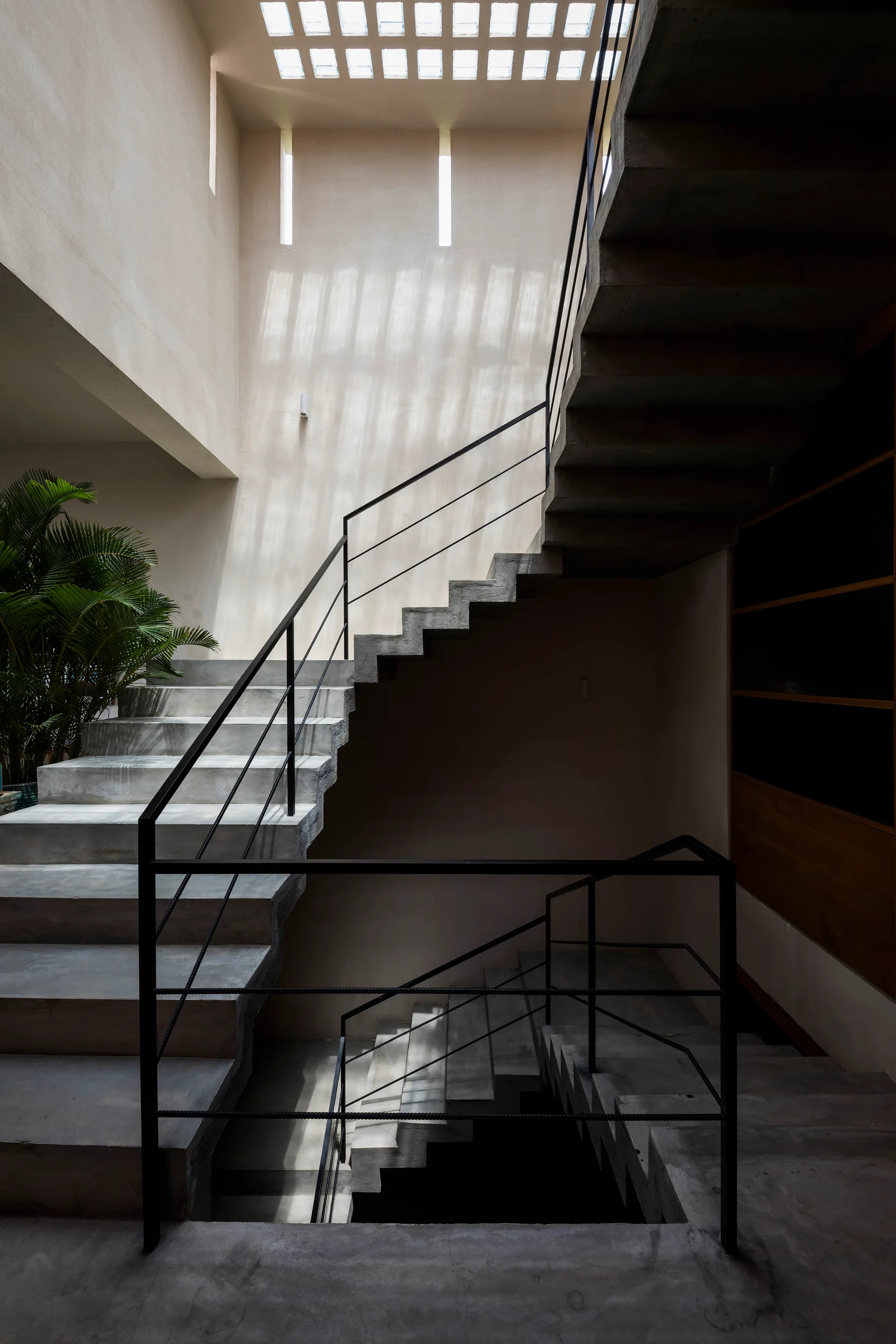 A multi-level raw concrete staircase with thin black metal railings winds upward under a geometric grid skylight in a minimalist beige stairwell.