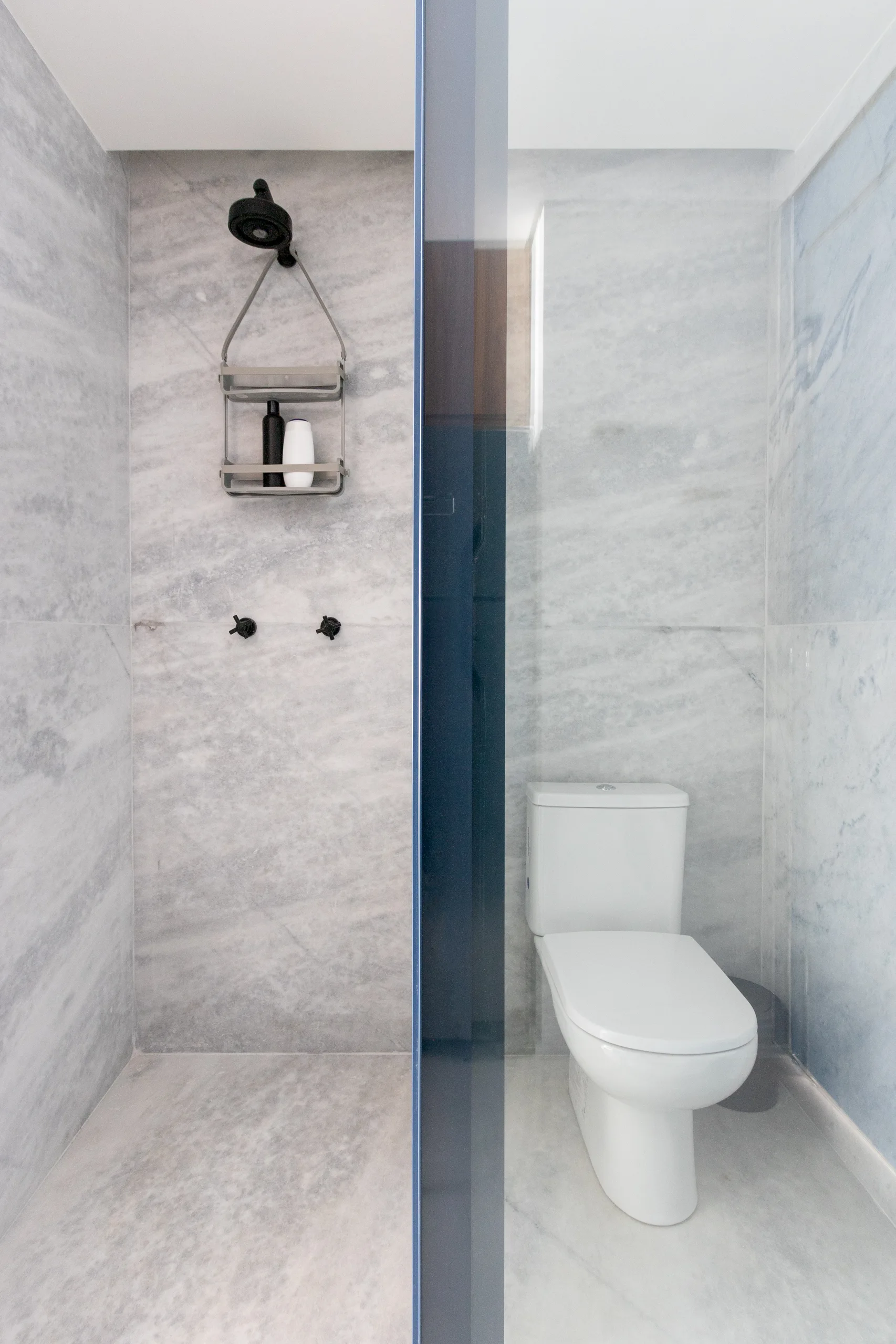 A modern bathroom with grey marble tiled walls features a white toilet next to a shower area with a dark blue tinted glass divider.