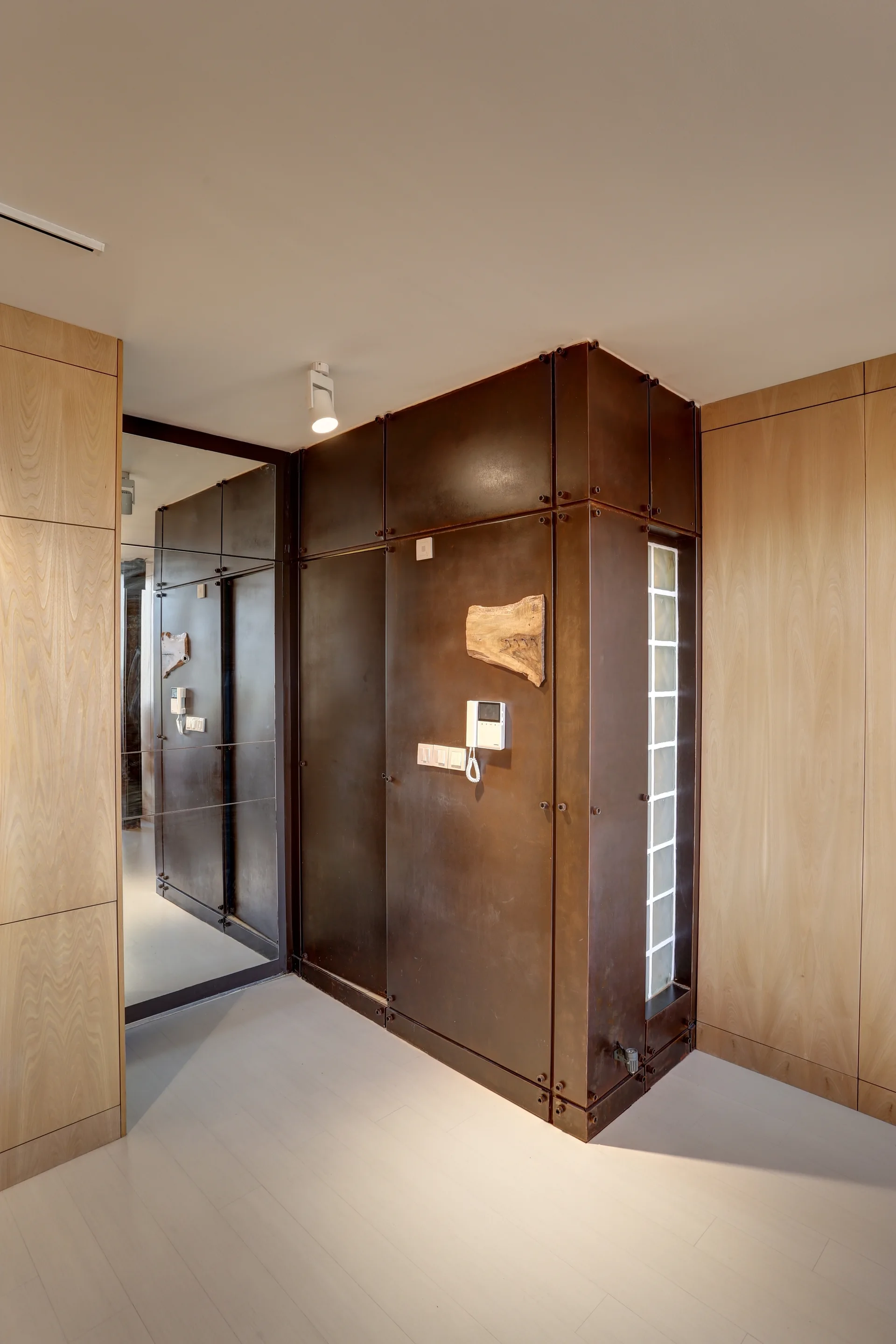 An entryway hallway features dark, bolted metal wall panels next to a vertical strip of glass blocks, light wood cabinetry, and a mirrored wall.