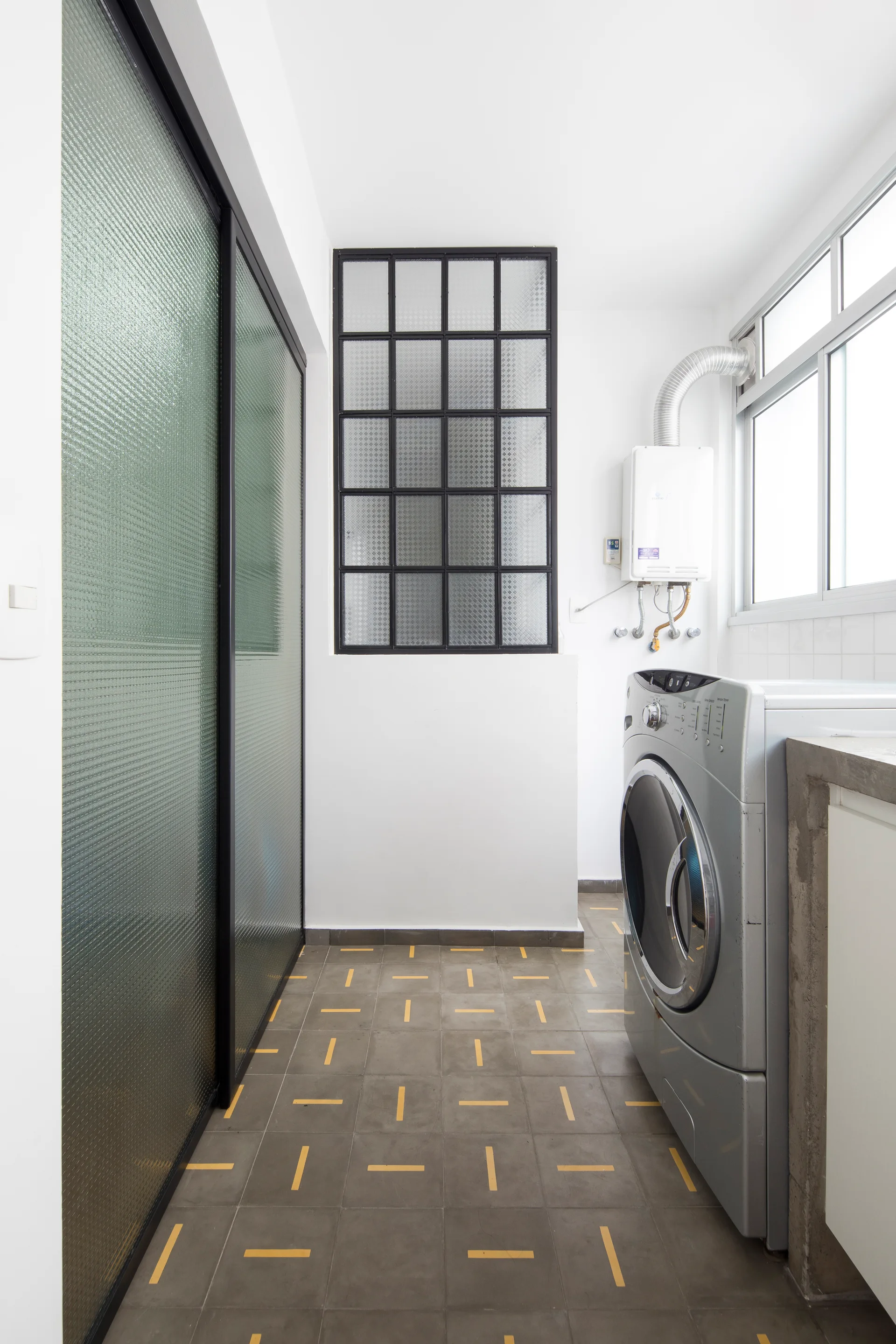 A utility laundry room features a silver washing machine next to a concrete counter, with textured glass sliding doors and a black grid window.