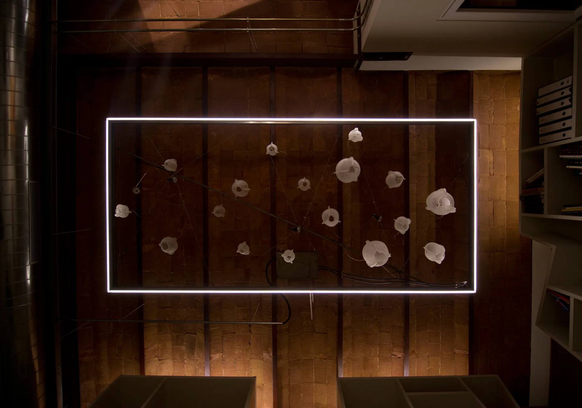 An overhead view of a brick ceiling with dark wood beams, a metal duct, and a glowing rectangular frame enclosing several small white pendant lamps.