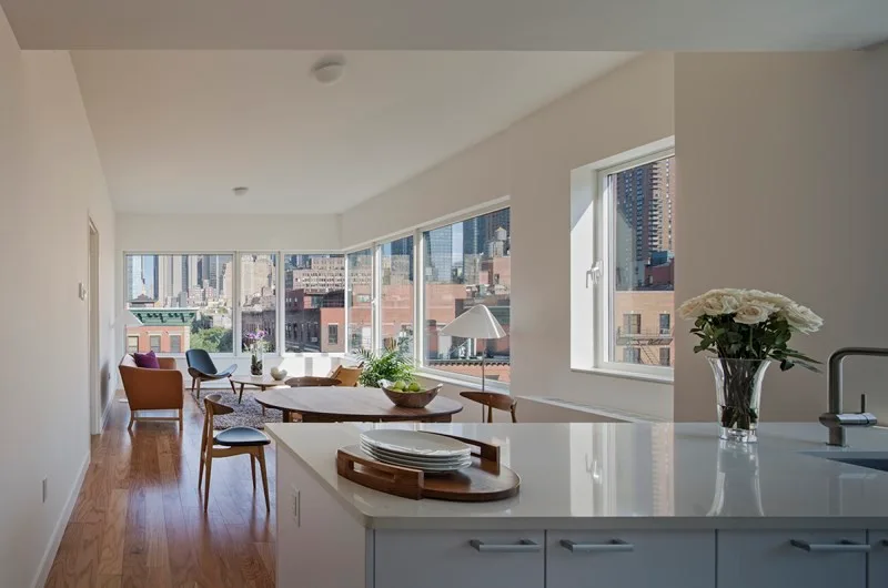 An open-concept apartment interior featuring a white kitchen island in the foreground, wood dining furniture, and large windows with a city view.