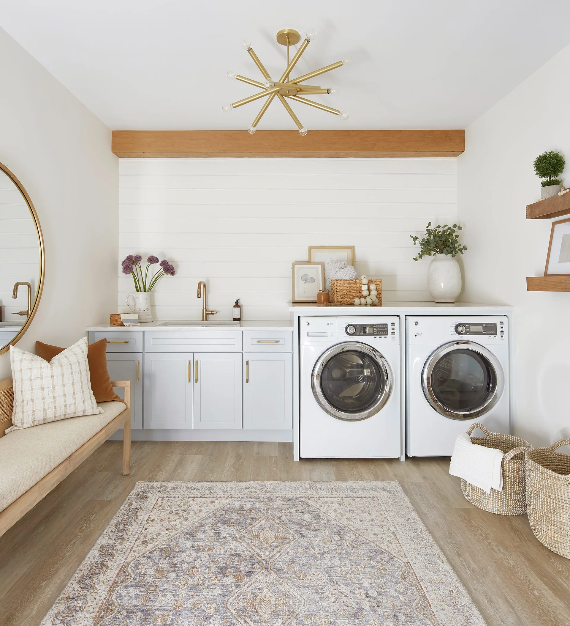 A bright laundry room features light blue cabinetry with gold hardware, a white shiplap wall, a wooden ceiling beam, and side-by-side laundry machines.