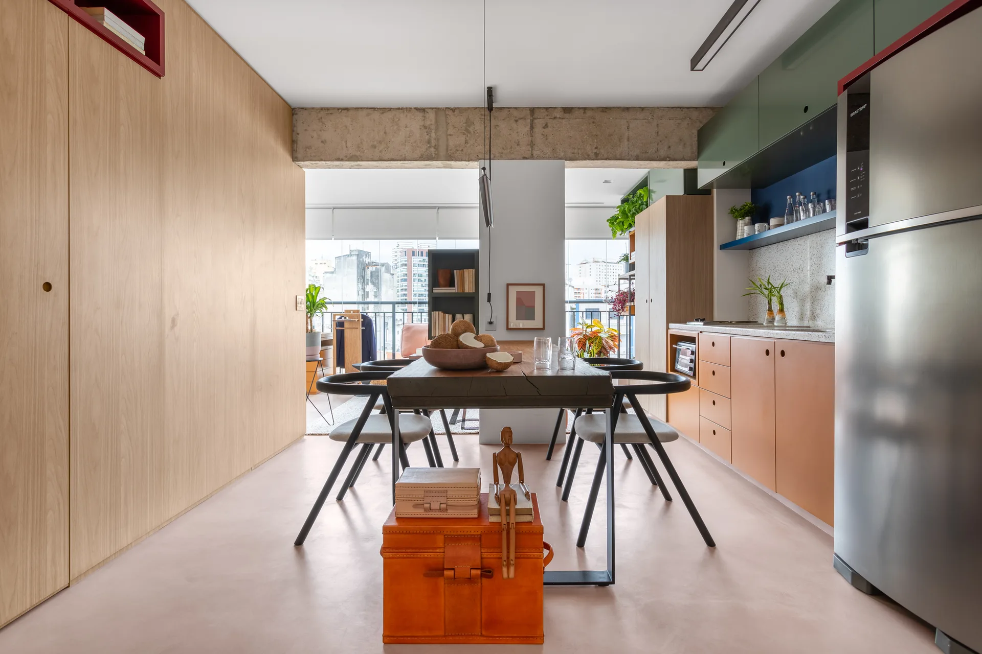 An open-plan dining space features a wooden table with black chairs beneath an exposed concrete beam, adjacent to a kitchen with wood and green cabinets.