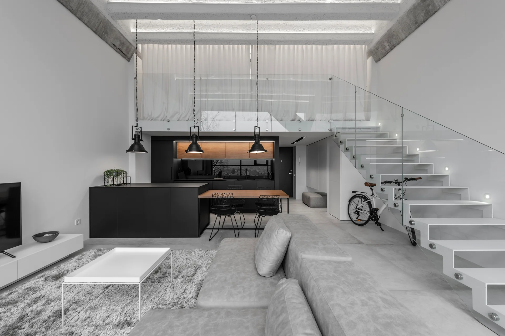 A modern open-plan living room with a grey sectional sofa, white floating staircase with glass railing, and a black kitchen island.