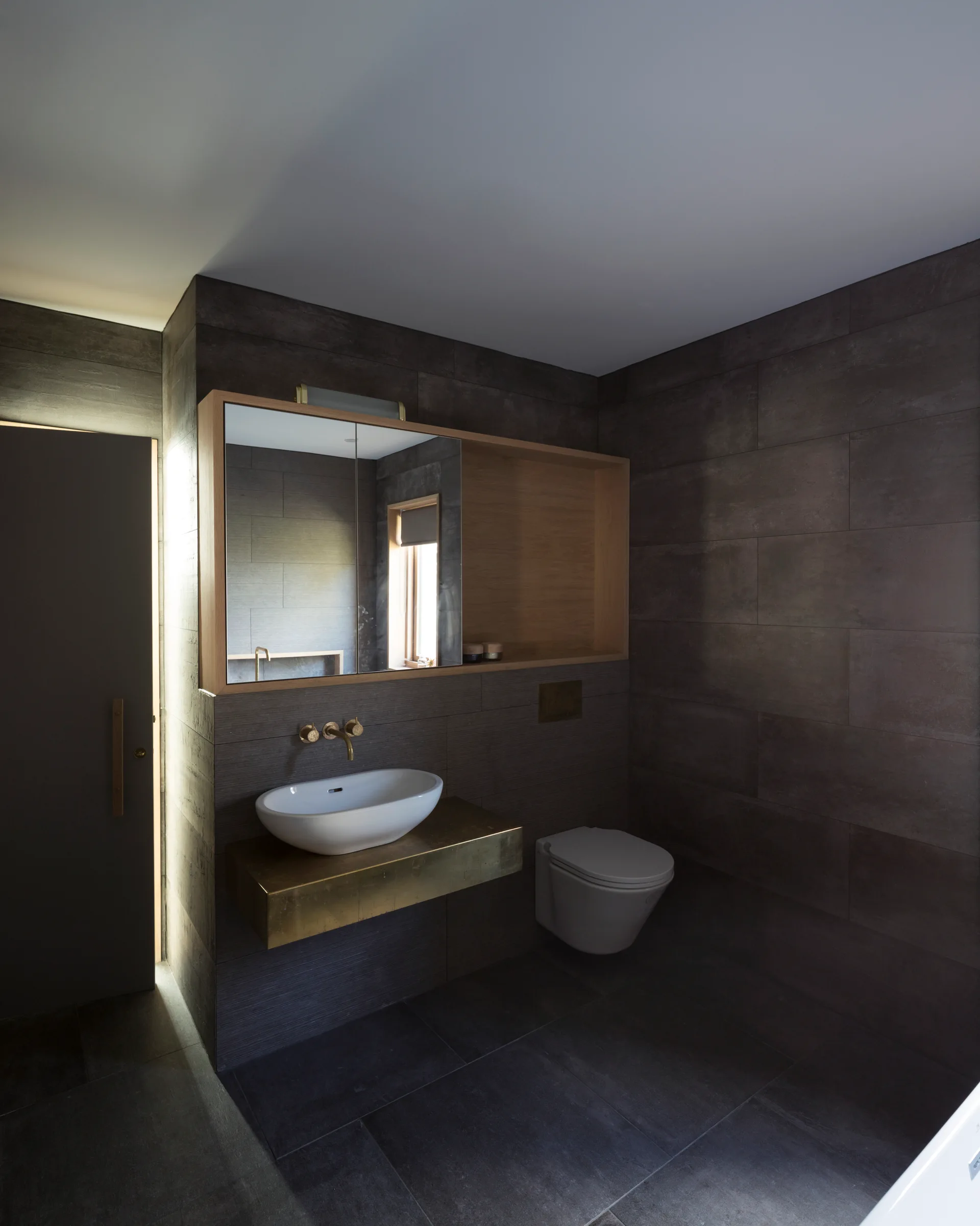 A modern bathroom featuring dark charcoal wall tiles, a wall-hung white toilet, and a white vessel sink resting on a gold brass floating vanity shelf.