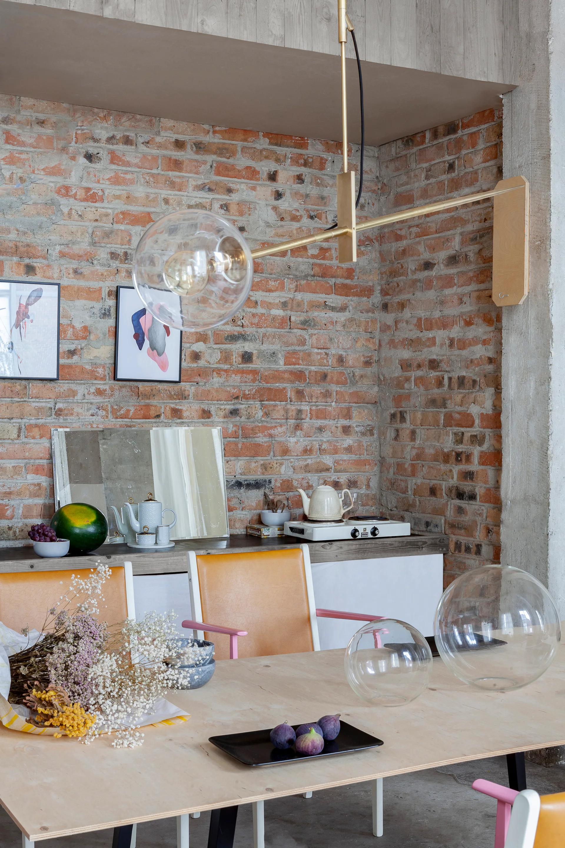 An eclectic dining room featuring a light wood table and yellow chairs set against an exposed brick wall with a brass pendant light.
