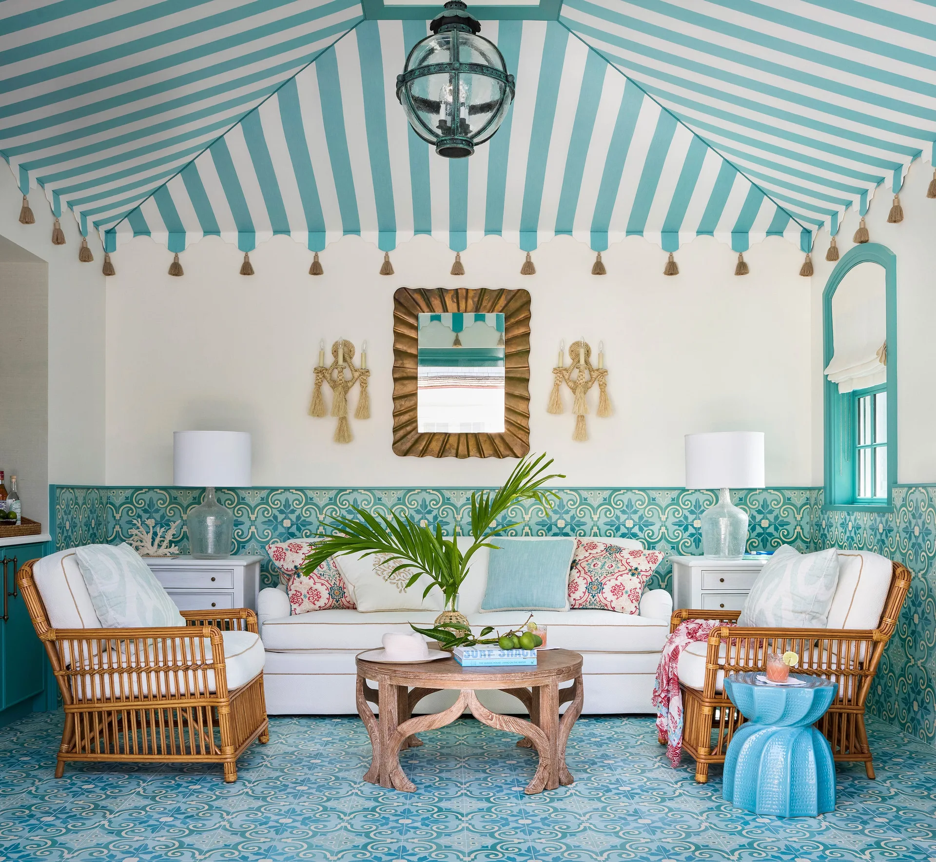 A living room features a blue and white striped tented ceiling, patterned teal wall and floor tiles, a white sofa, and rattan armchairs.