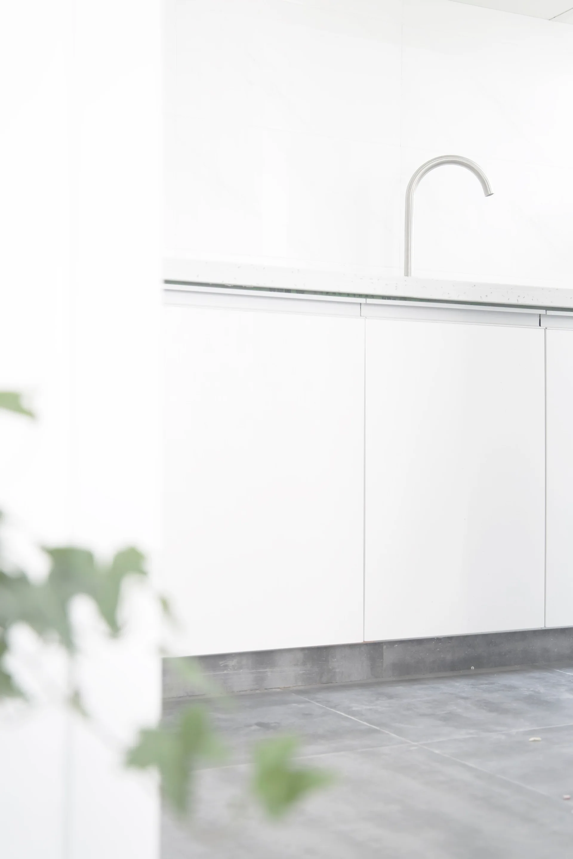 A close-up of minimalist white handleless kitchen cabinets, a light stone countertop with a curved metal faucet, and grey tiled floors.