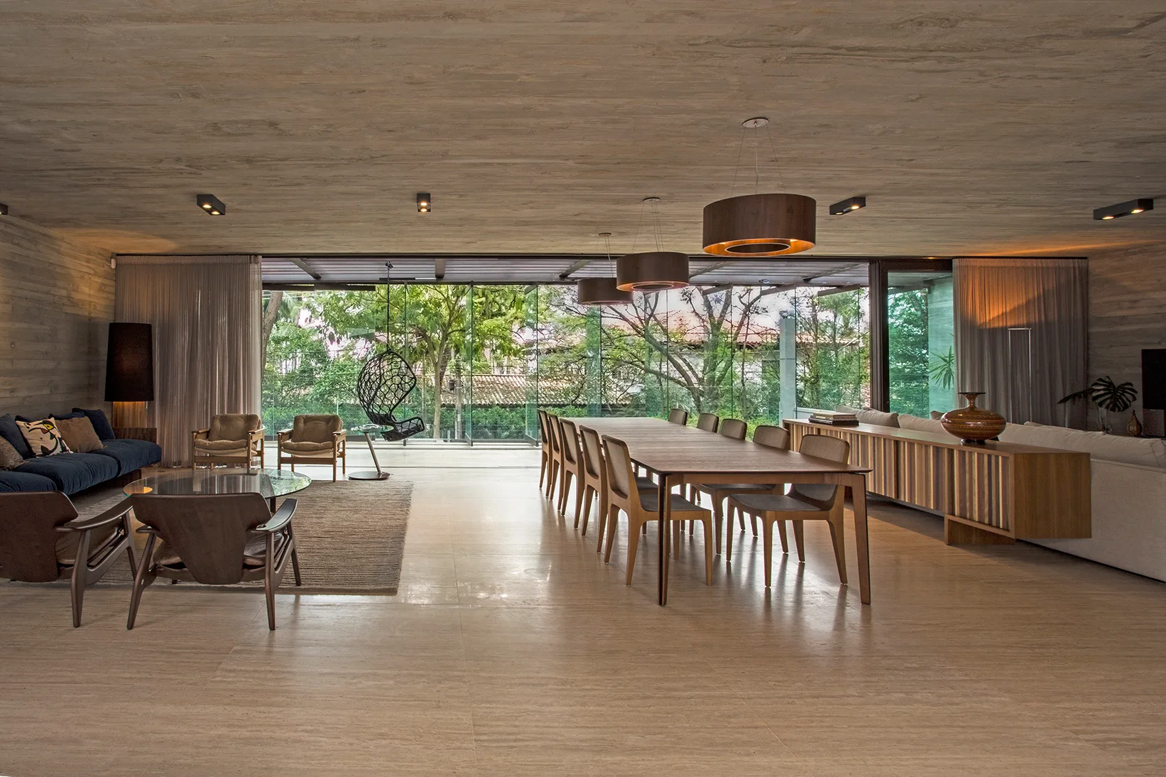 A large open-concept living and dining space with a board-formed concrete ceiling, a long wooden dining table, and floor-to-ceiling glass doors.