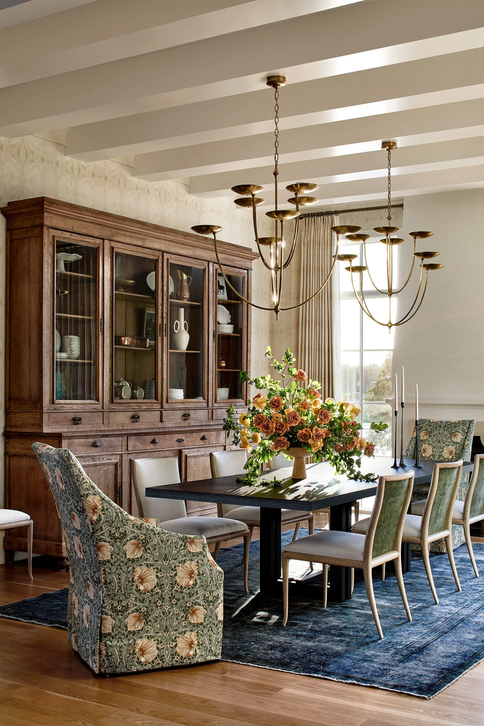 A formal dining room features a dark table, patterned upholstered chairs, a large wooden cabinet, and twin brass chandeliers hanging from a beamed ceiling.