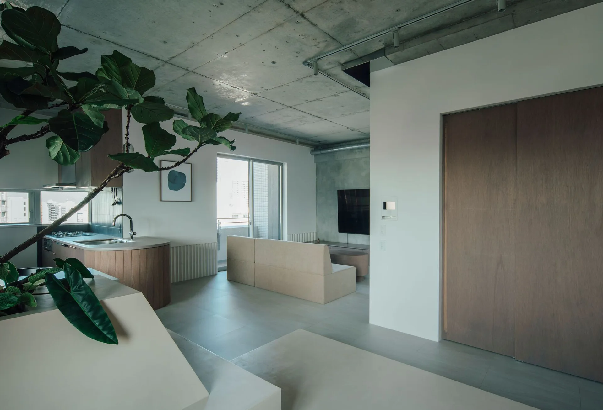 An open-concept living area features an exposed concrete ceiling, a low-profile beige sofa, and a kitchen island with a curved wood base.