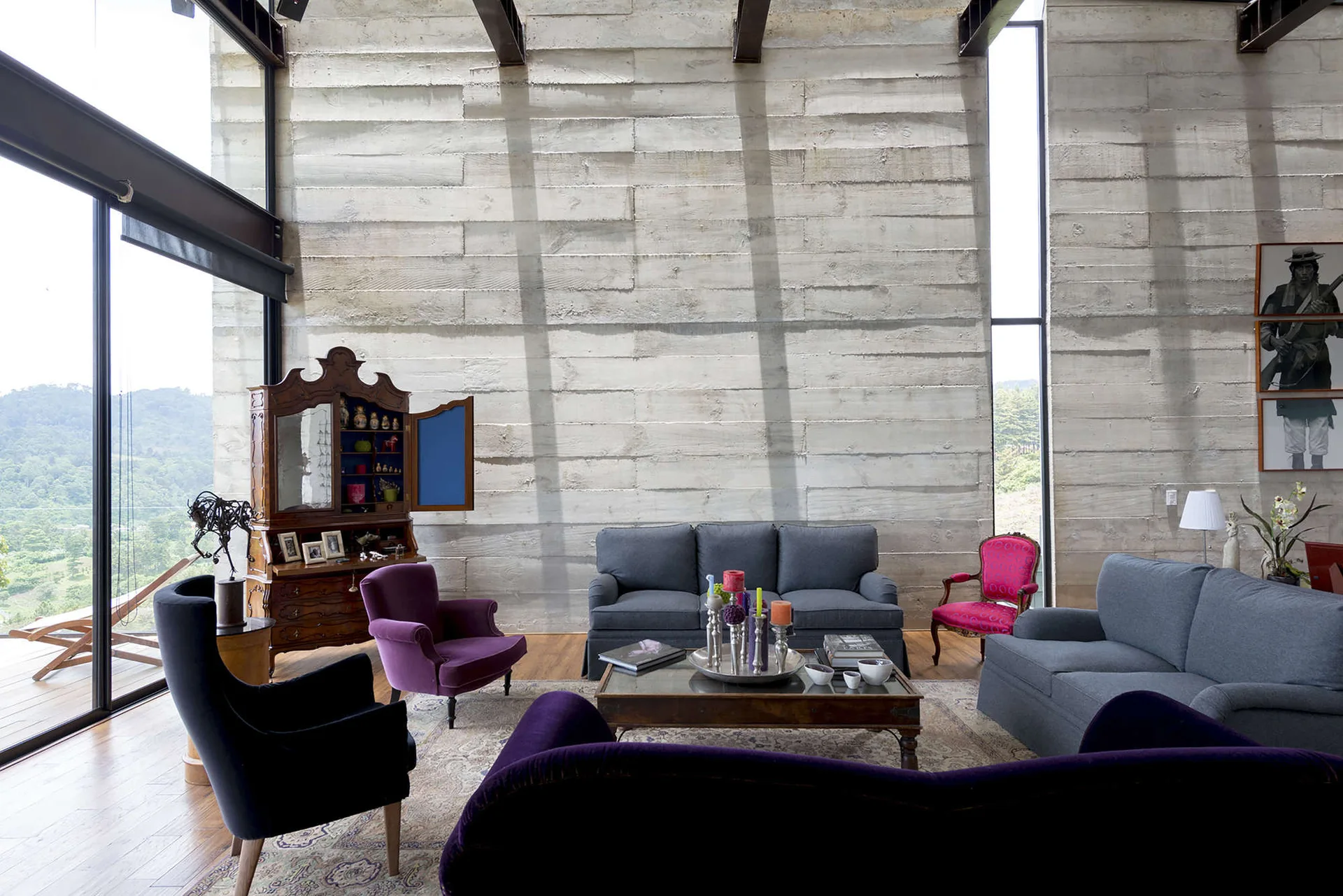 An eclectic living room featuring a massive board-formed concrete wall, gray sofas, purple velvet armchairs, and floor-to-ceiling windows.