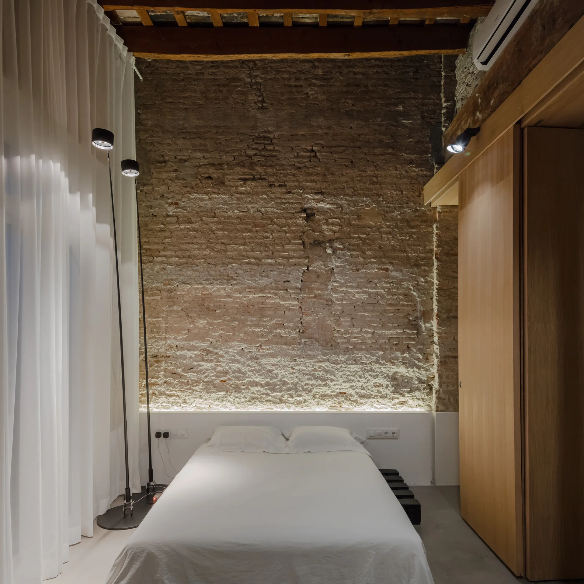 A low white bed sits in front of a tall exposed brick wall illuminated by a soft LED light strip in an industrial style bedroom.