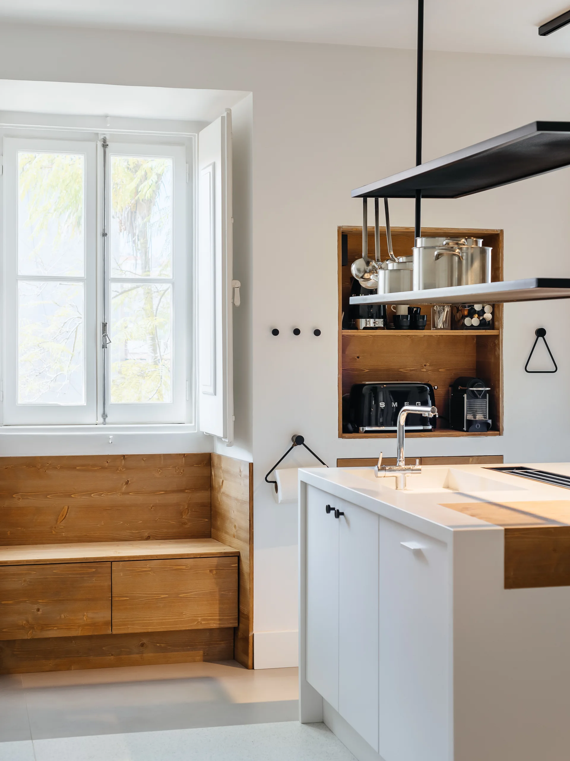 A minimalist white kitchen featuring a kitchen island, a built-in wooden bench below a window with shutters, and recessed wooden wall shelving.