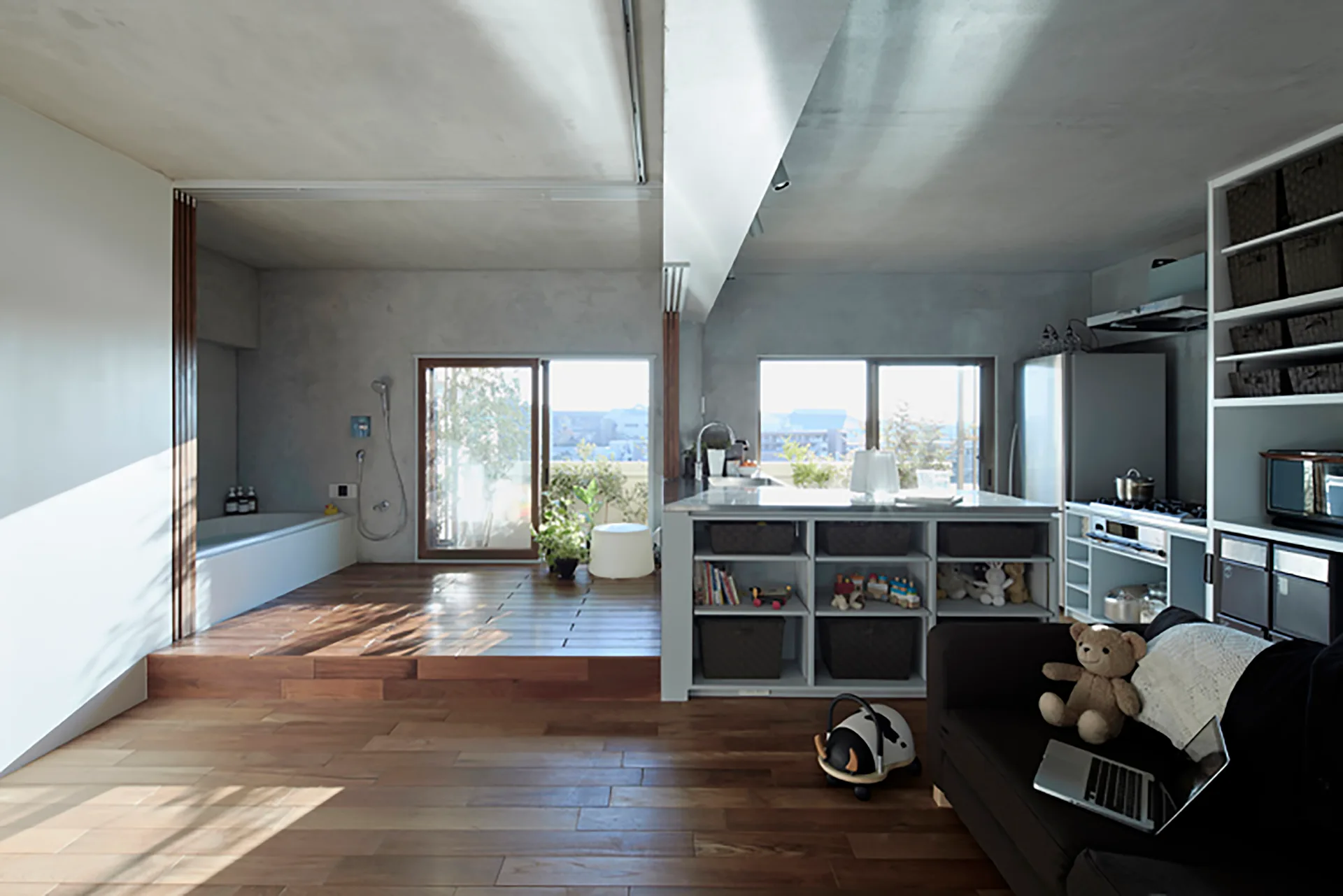 An open-concept home features a kitchen island with storage baskets next to a raised wooden platform with a built-in bathtub and glass sliding doors.