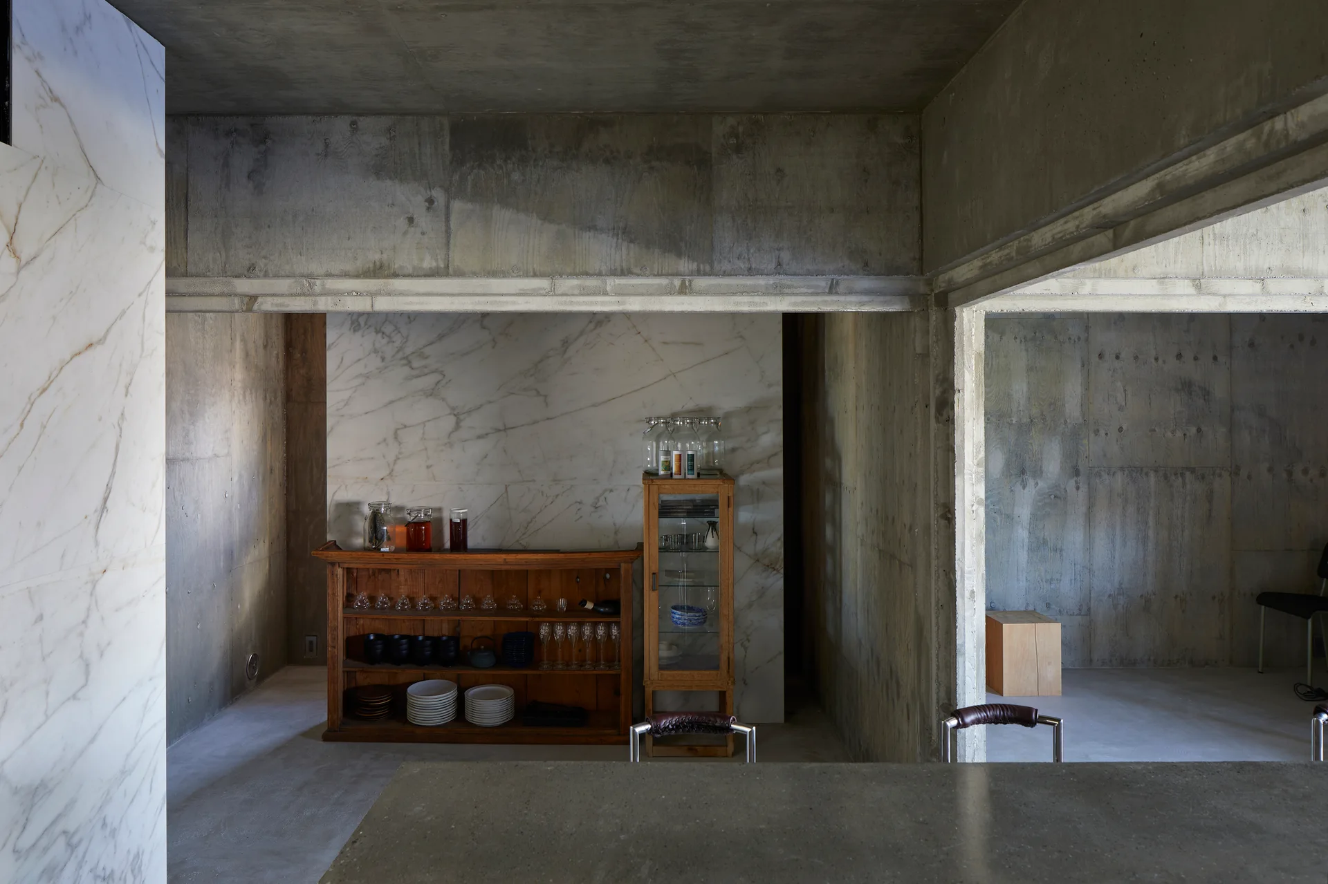 A minimalist kitchen features raw concrete walls and ceiling beams, a white marble accent wall, and freestanding wooden cabinetry holding glassware.