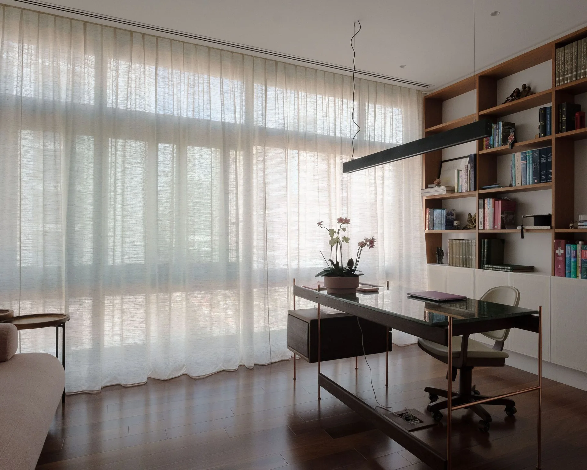A contemporary home office with a glass-topped desk, a built-in wooden bookshelf, and full-length sheer curtains filtering natural light.