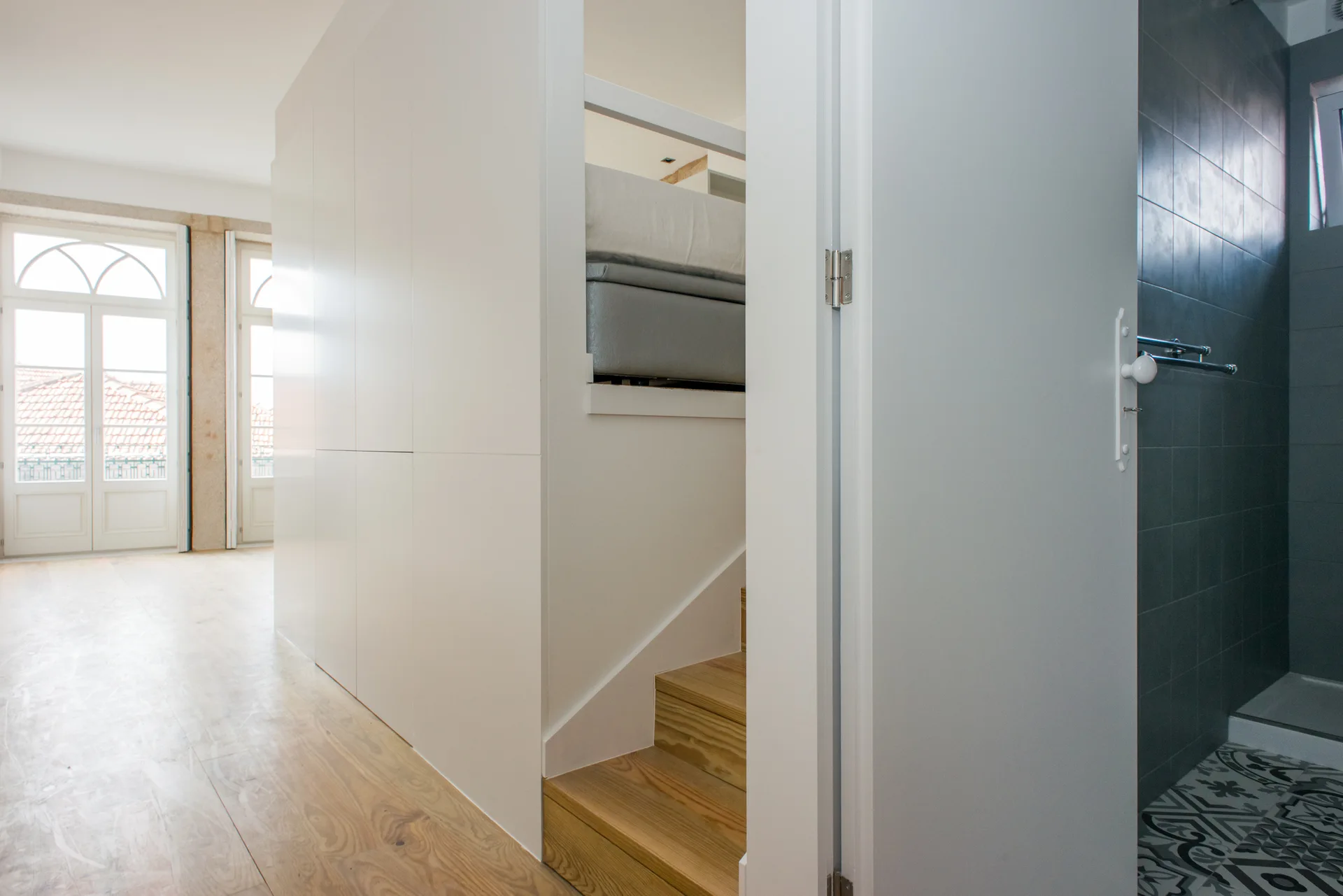 A minimalist room featuring a white built-in storage unit with integrated wooden steps leading to a loft bed next to a tiled bathroom doorway.