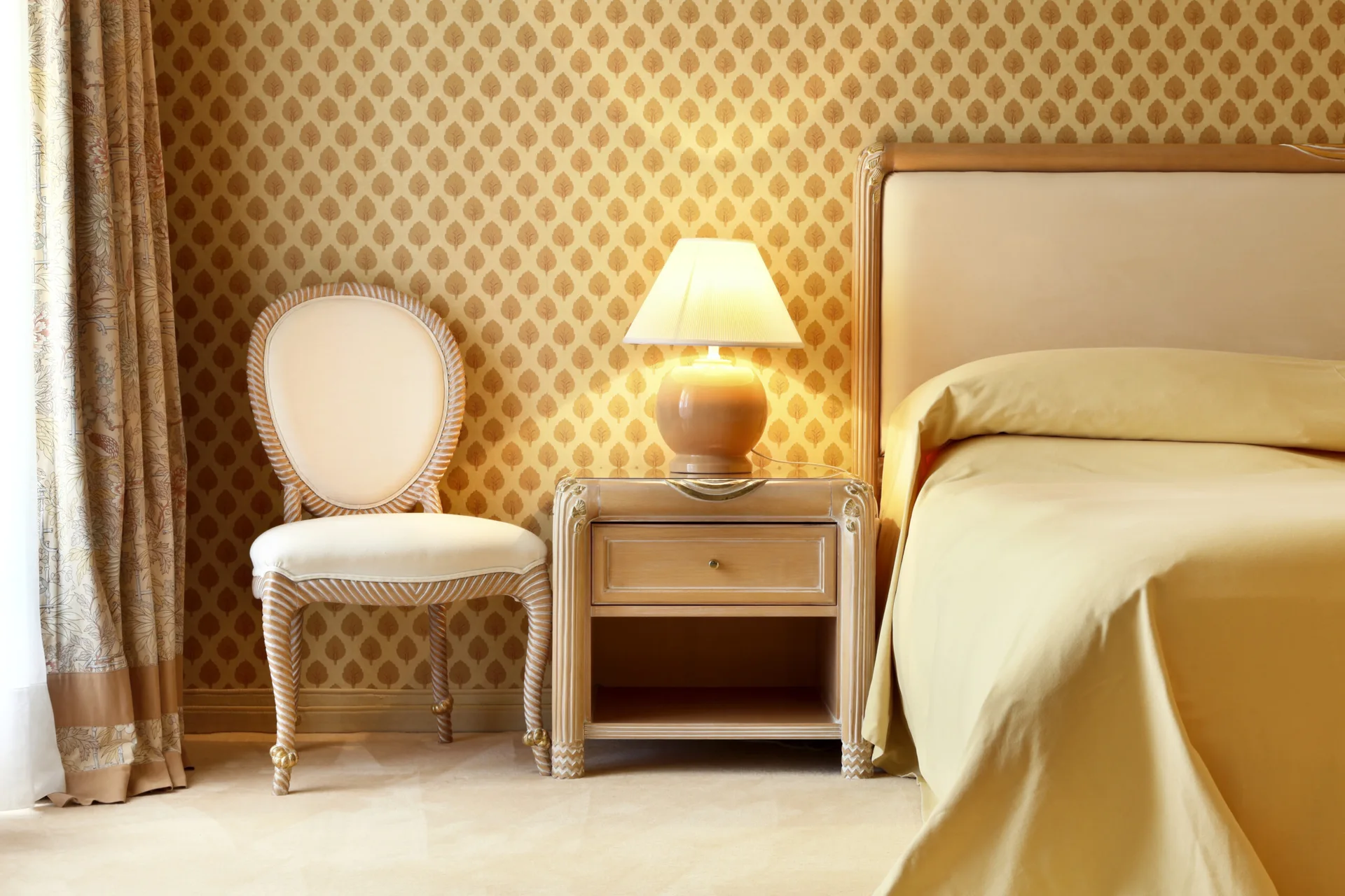 A classic bedroom features a bed with yellow linens, a wood nightstand with a lit lamp, and an oval-backed chair against leaf-patterned wallpaper.