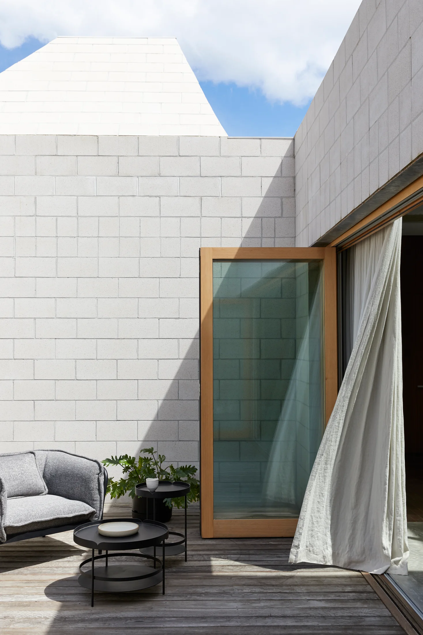 A minimalist wood deck patio features a grey armchair, black metal side tables, concrete block walls, and a large wood-framed glass sliding door.