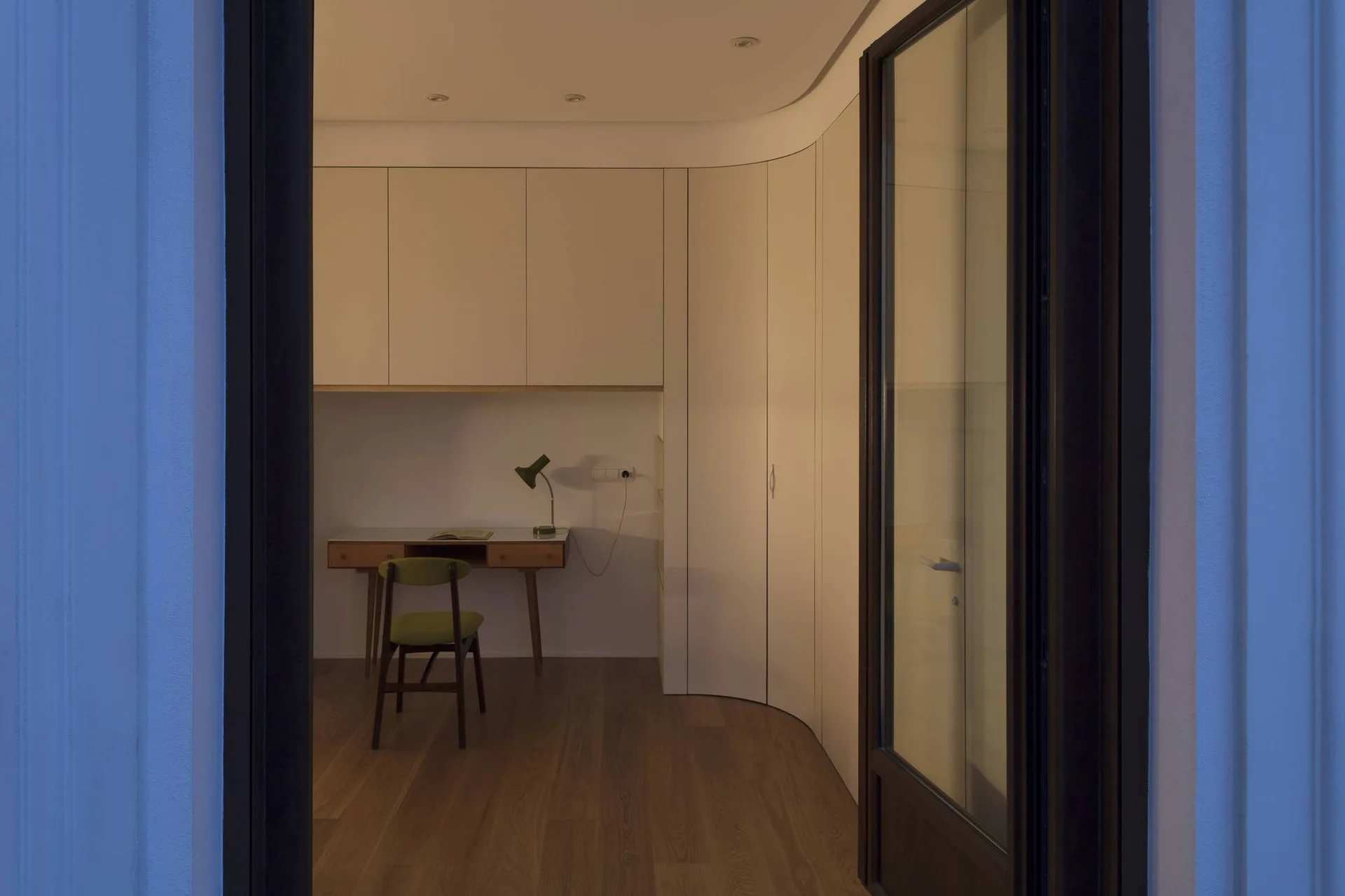 A minimalist home office viewed through a dark doorway, featuring a simple wooden desk, green chair, and curved off-white built-in cabinets.