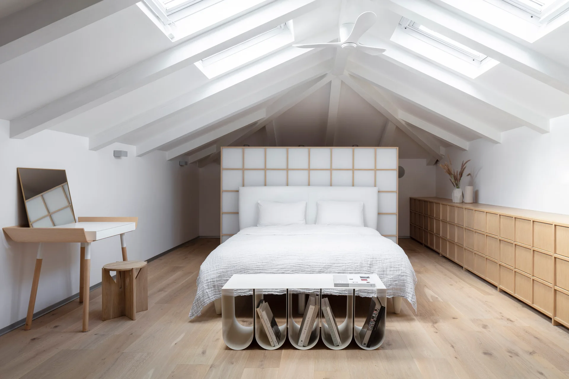 A minimalist attic bedroom features a bed with a grid headboard, a sloped ceiling with skylights, and light wood storage units.