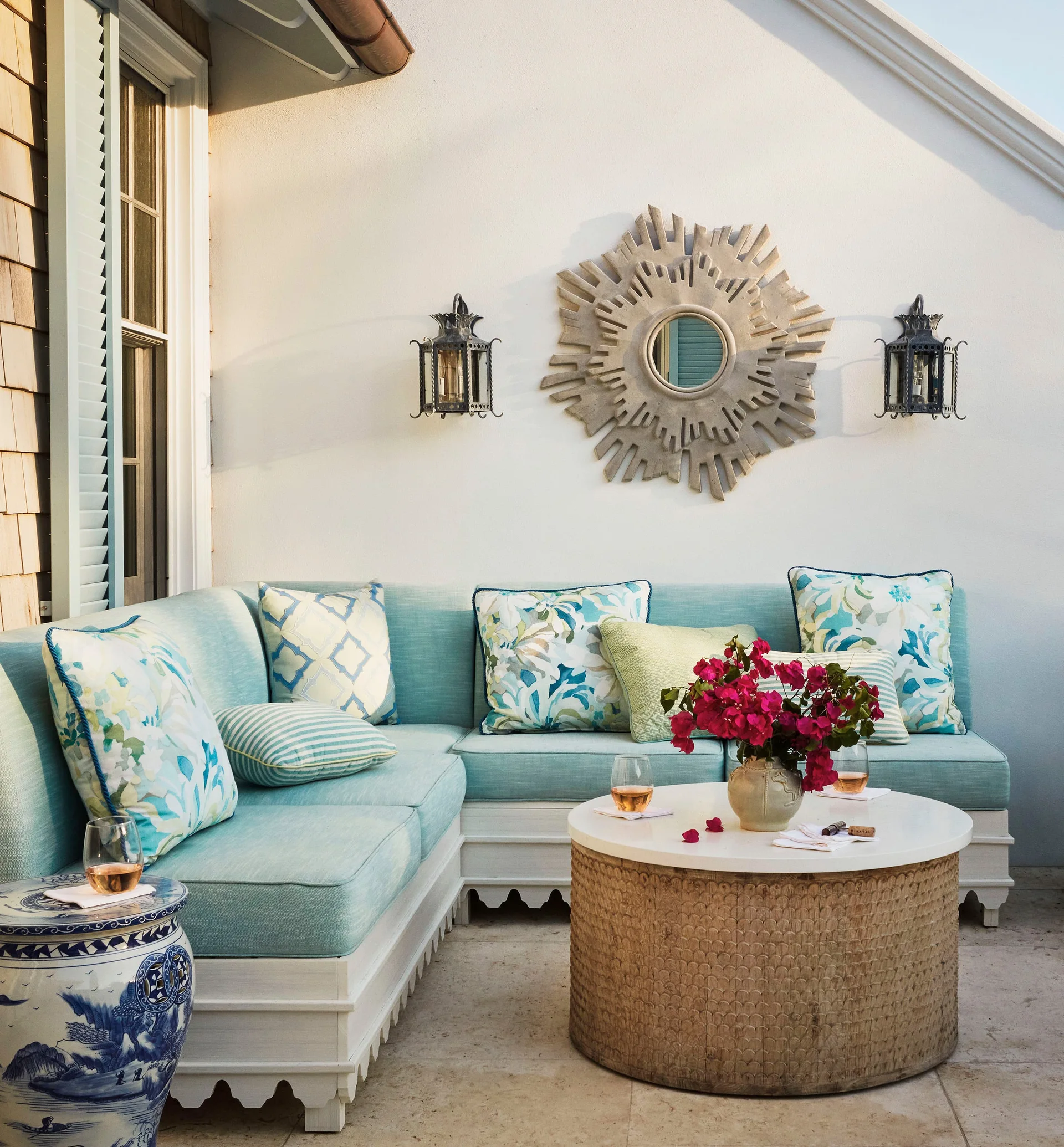 A light blue outdoor sectional sofa with patterned throw pillows sits on a stone patio next to a woven round coffee table and a sunburst wall mirror.