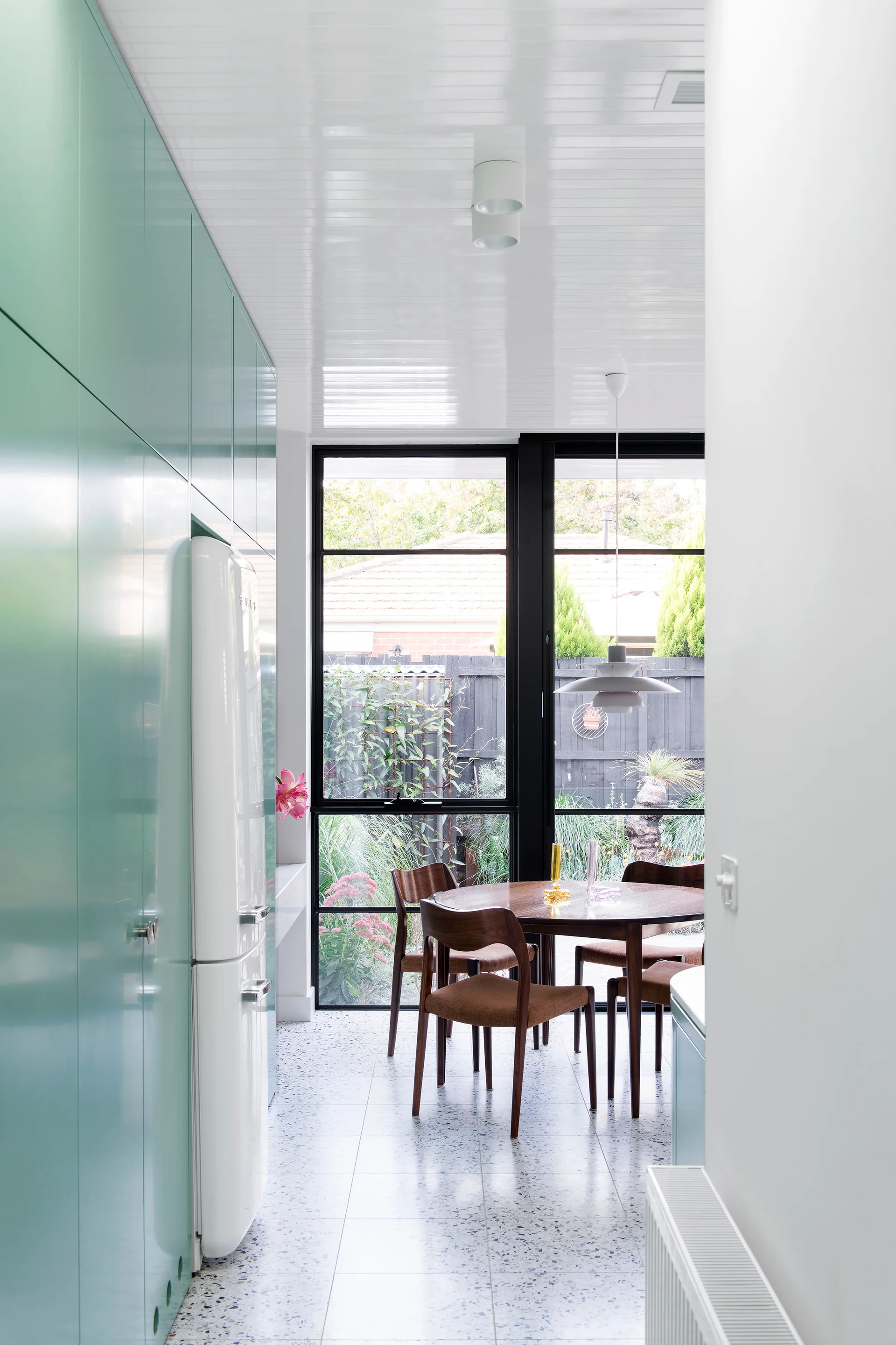 A modern dining area features a round wooden table, mint green cabinetry, terrazzo tile flooring, and a large black-framed window looking out.