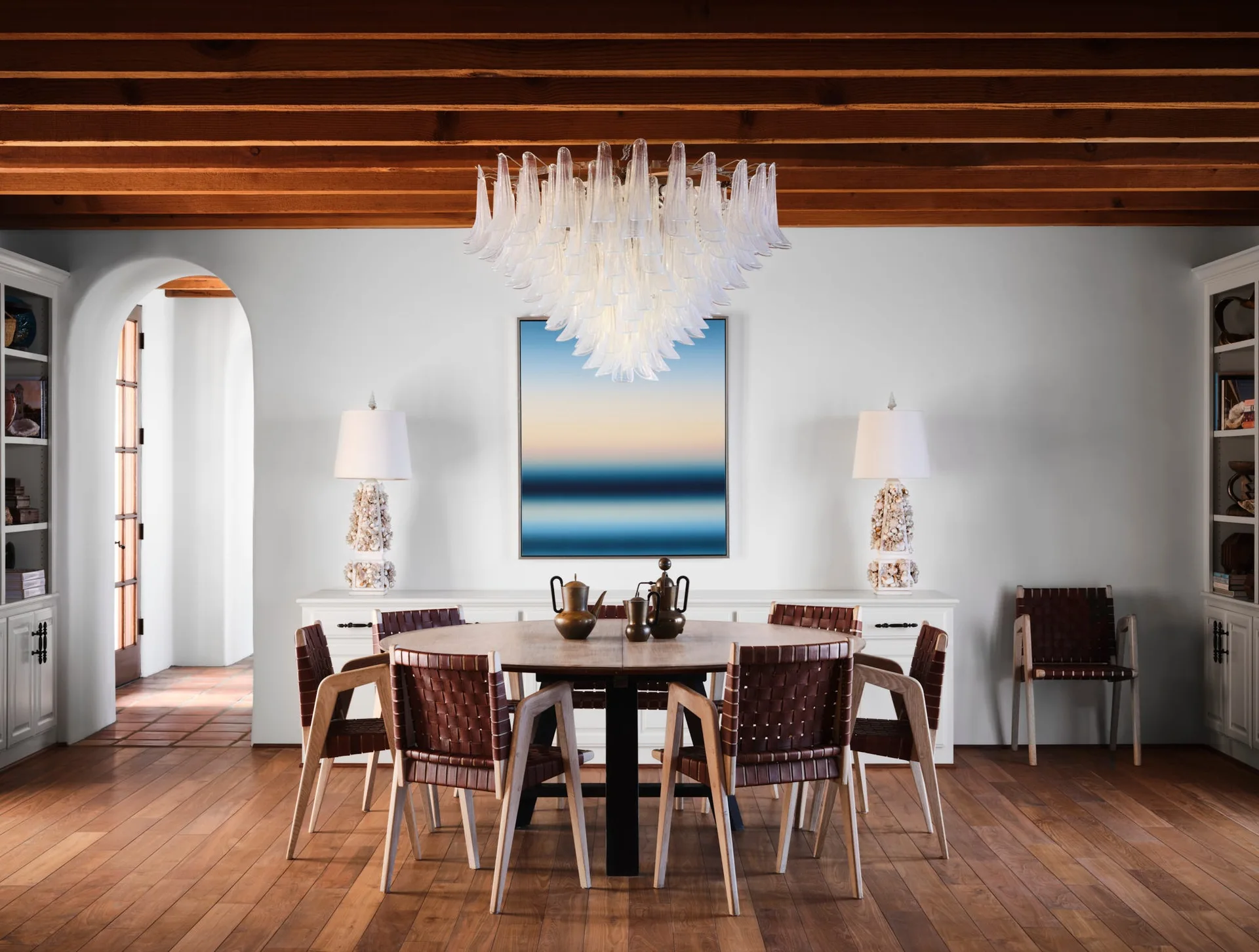 A dining room featuring a round wooden table with woven leather chairs, a large glass chandelier, and exposed wood ceiling beams.