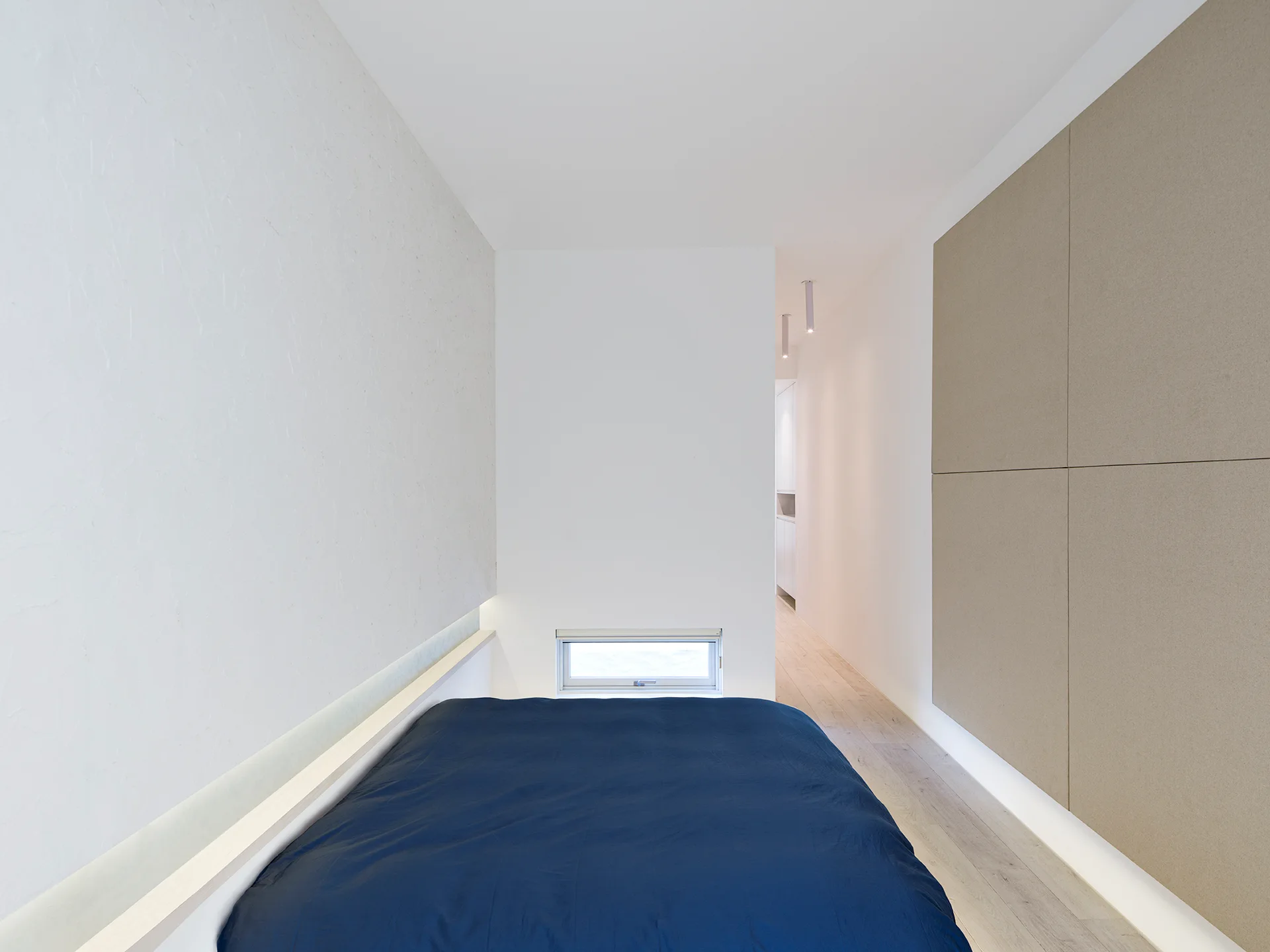A minimalist bedroom featuring a bed with a dark blue cover, a low-set horizontal window, and large square tan wall panels on a light wood floor.