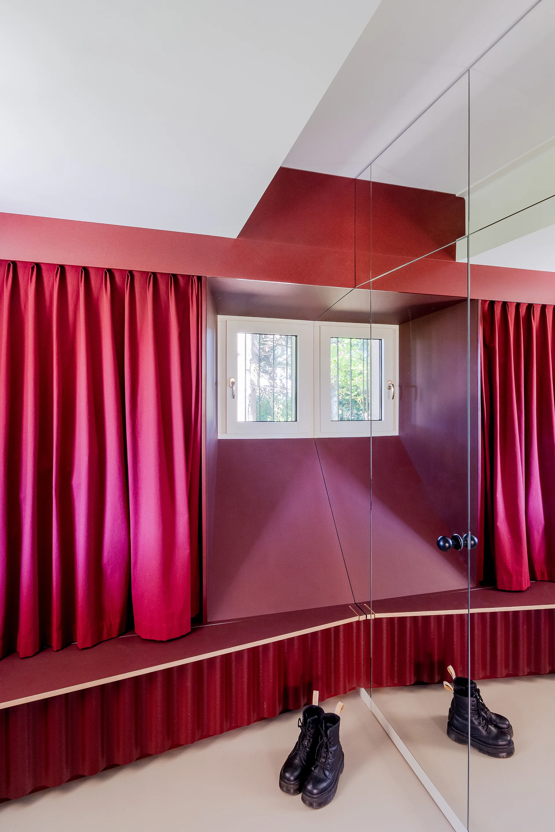 A deep red built-in bench with a corrugated front sits beneath a small window alongside matching red curtains and a large mirrored wall.