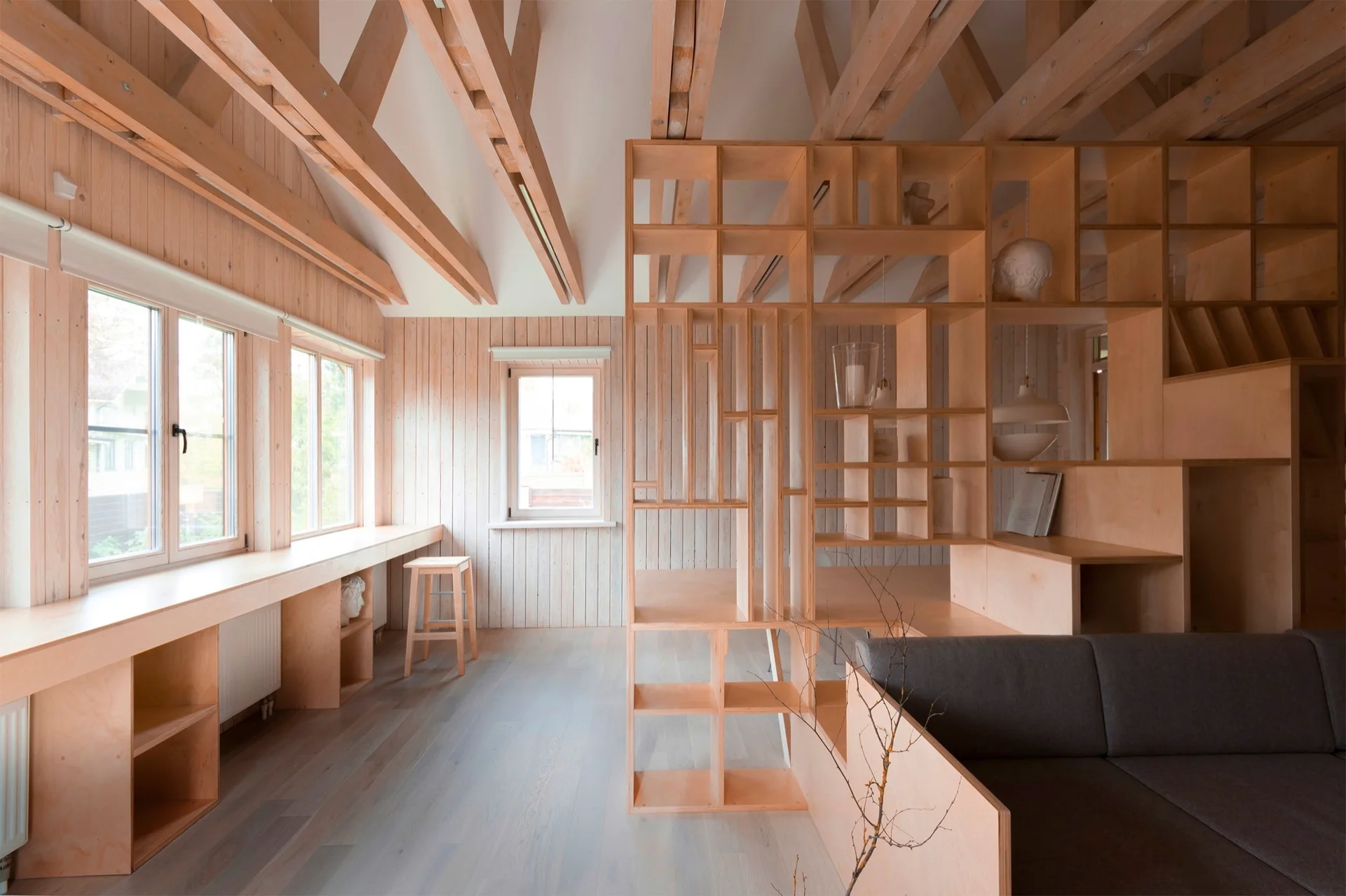 A light wood living room features a custom plywood room divider, exposed ceiling beams, vertical wall paneling, and windows.