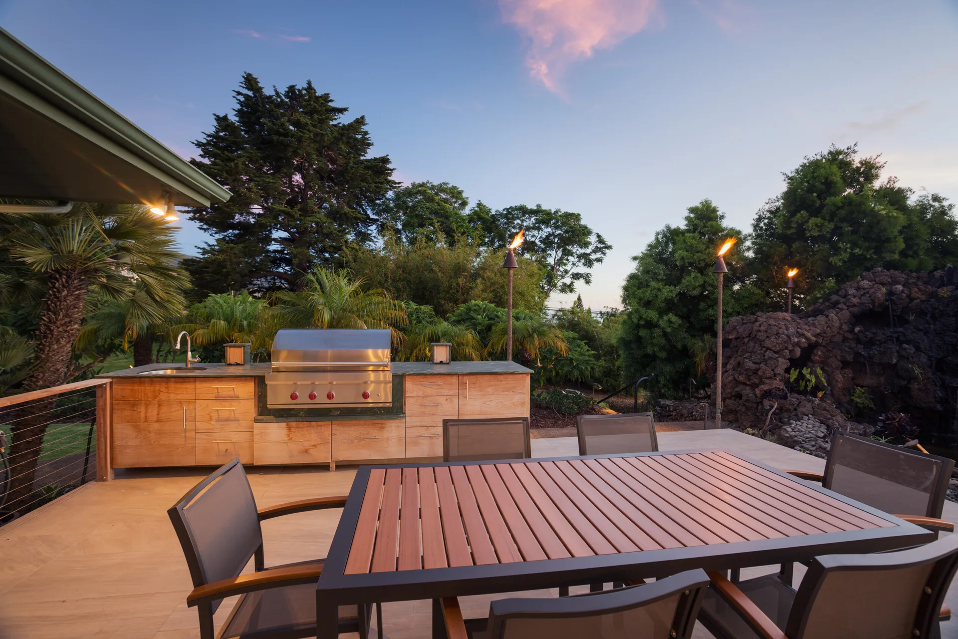 An outdoor kitchen with wood cabinets and a built-in grill sits on a stone tile patio next to a dining table and lush landscaping.