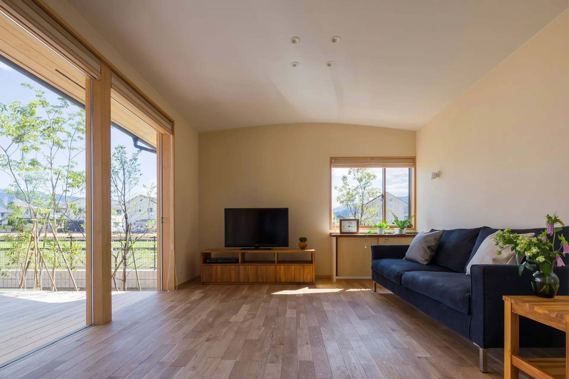 A minimalist living room features a dark blue sofa, a wooden TV stand, and large sliding glass doors opening to a bright outdoor deck.