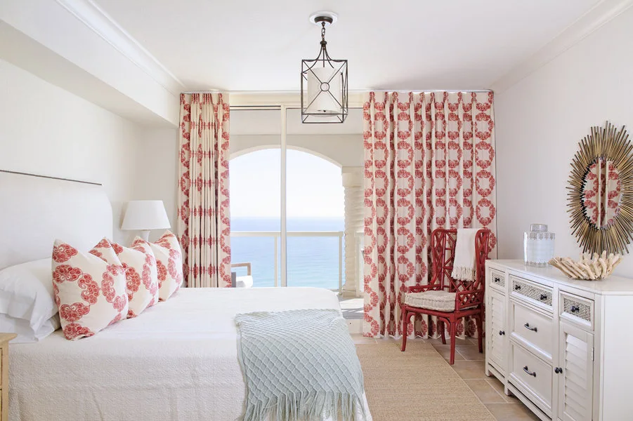 A bright coastal bedroom with red patterned curtains and pillows, a white bed, a louvered dresser, and glass doors opening to the ocean.