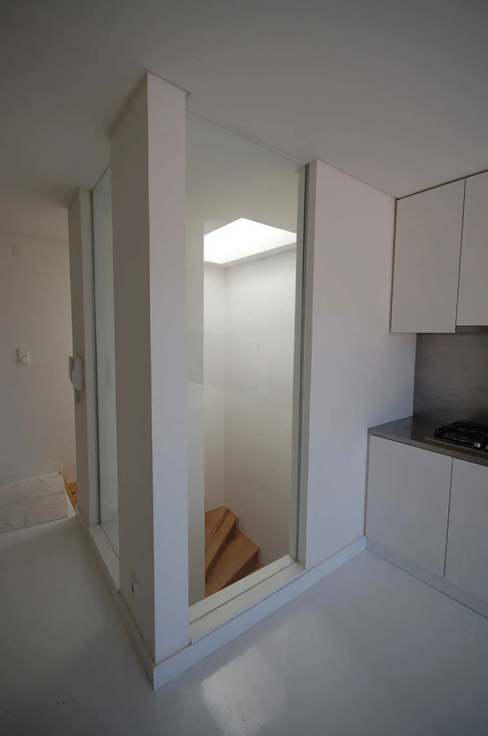 A minimalist white kitchen adjacent to a wooden staircase enclosed by a floor-to-ceiling glass partition under a skylight.