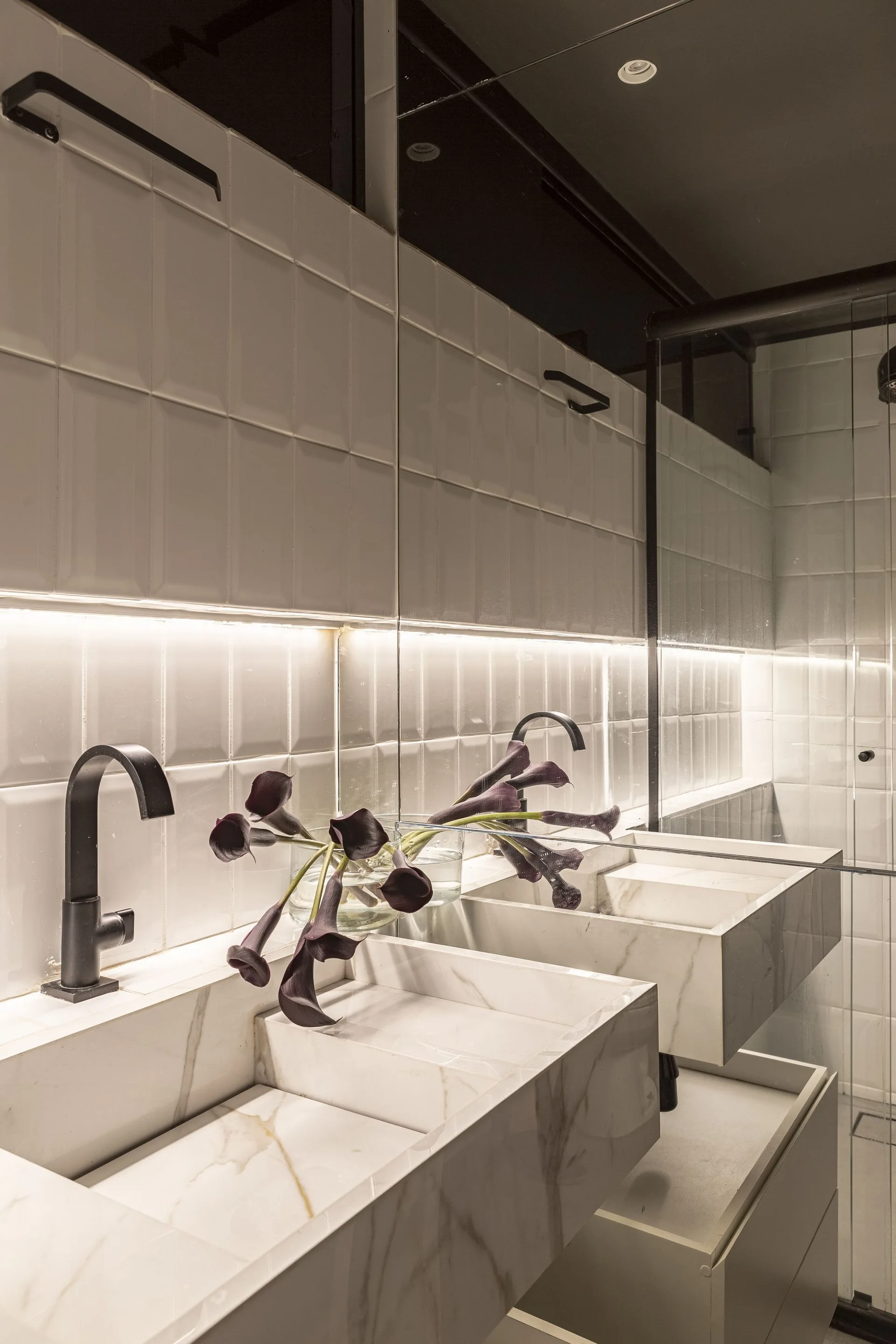 Two white marble slab sinks with matte black faucets sit beneath a mirrored cabinet lit by an under-shelf LED strip.