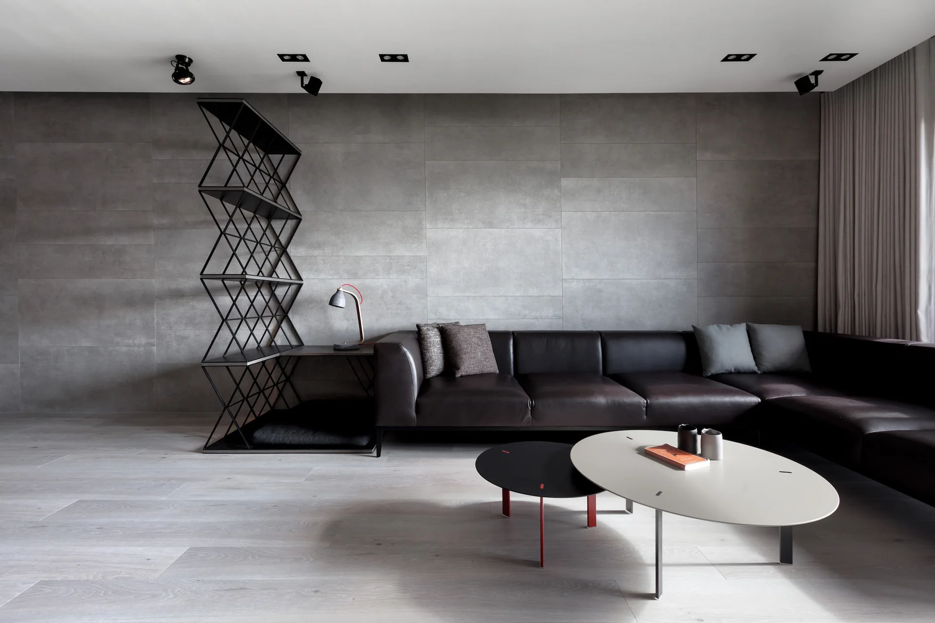 A minimalist living room with a dark leather sectional sofa, elliptical nesting tables, and a geometric metal bookshelf against a grey tile wall.