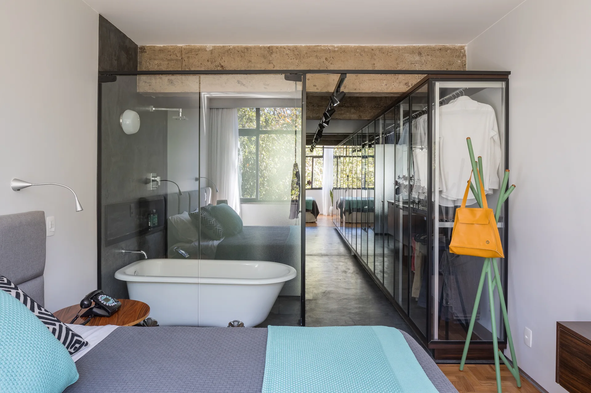 A modern bedroom features a freestanding bathtub inside a glass-walled enclosure, adjacent to a glass-front closet and concrete ceiling beams.