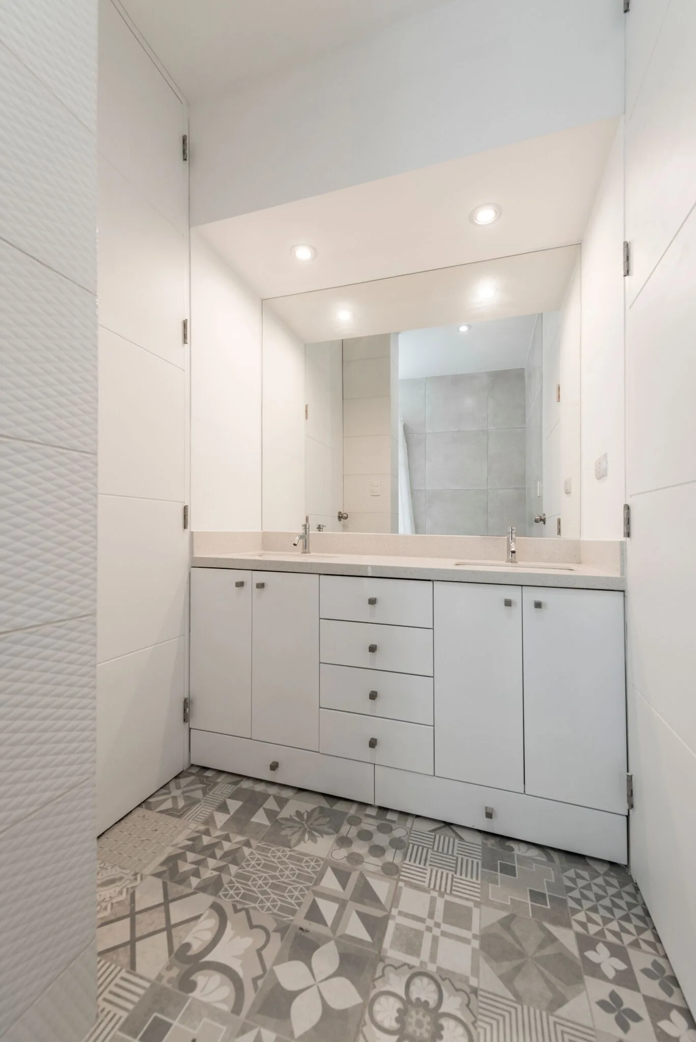 A minimalist white bathroom features a double sink vanity, a large wall-mounted mirror, and patterned grey and white floor tiles.
