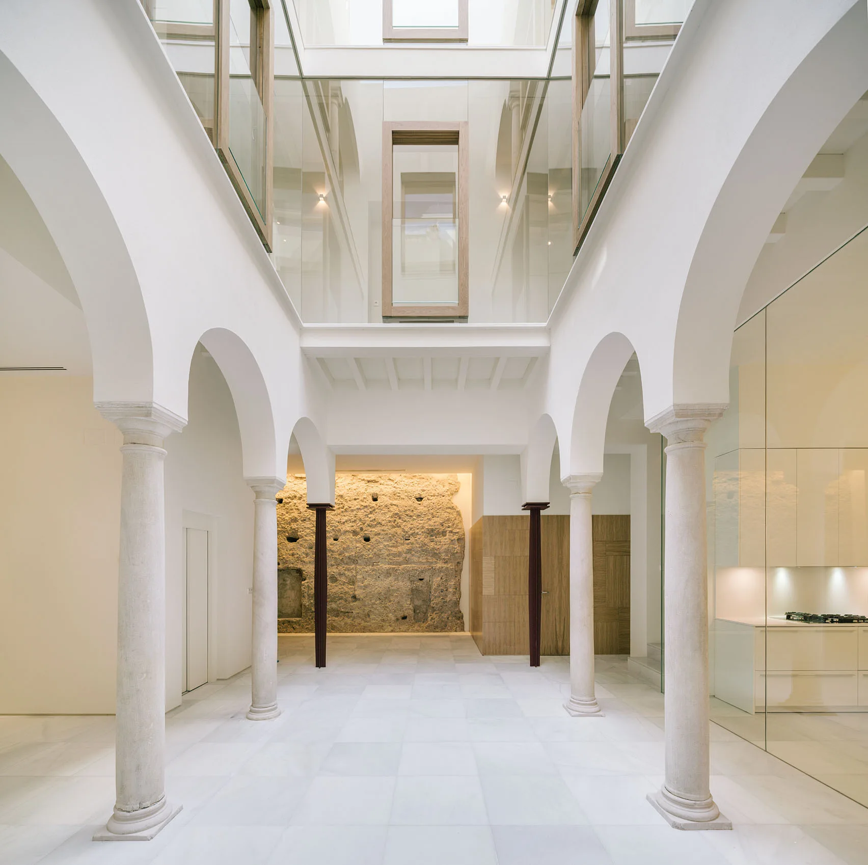 A bright white courtyard atrium features classical stone columns and arches beneath a glass ceiling and an exposed historical stone wall in the background.