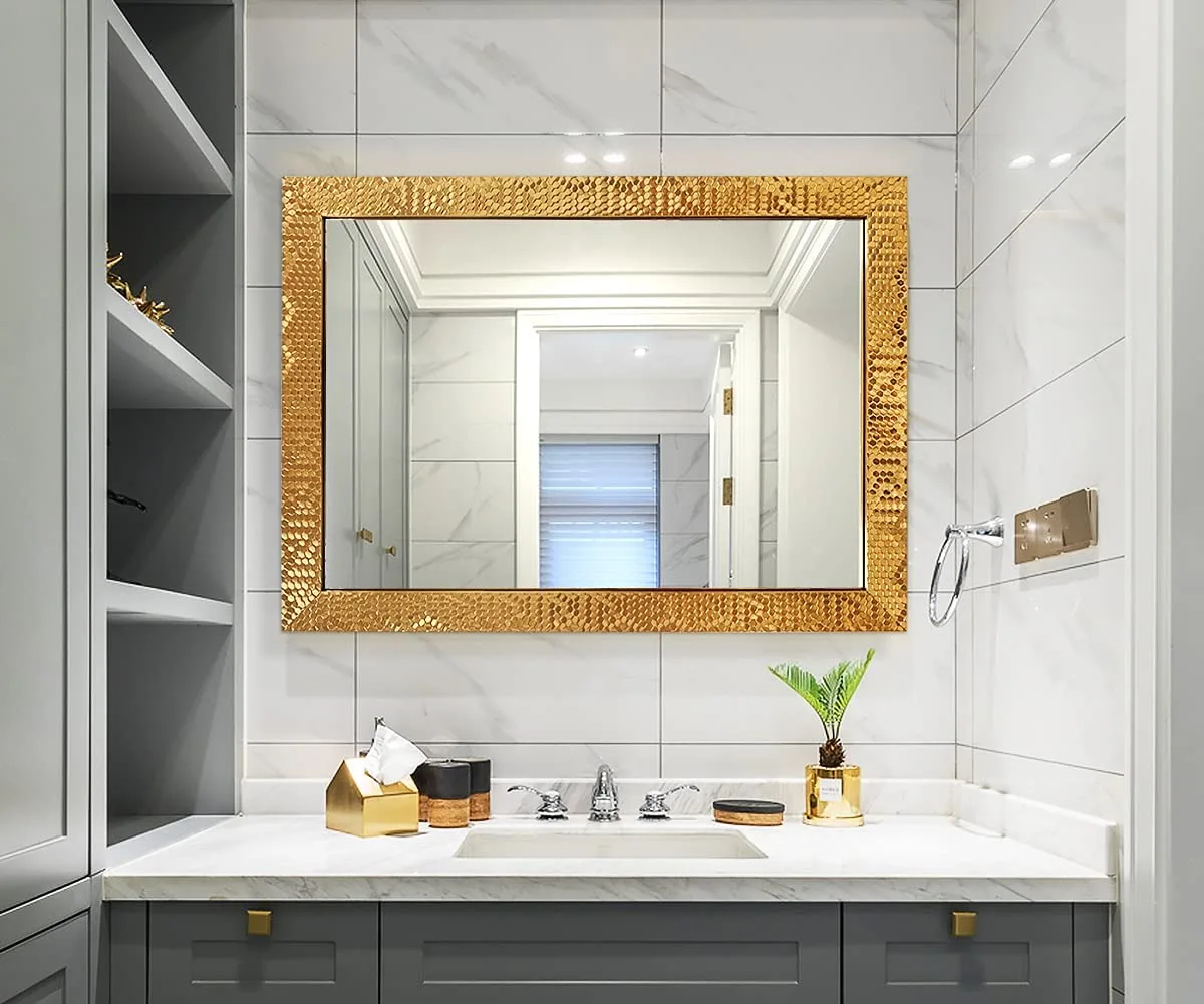 A grey bathroom vanity with a white marble countertop is set against marble wall tiles beneath a large rectangular mirror in a textured gold frame.