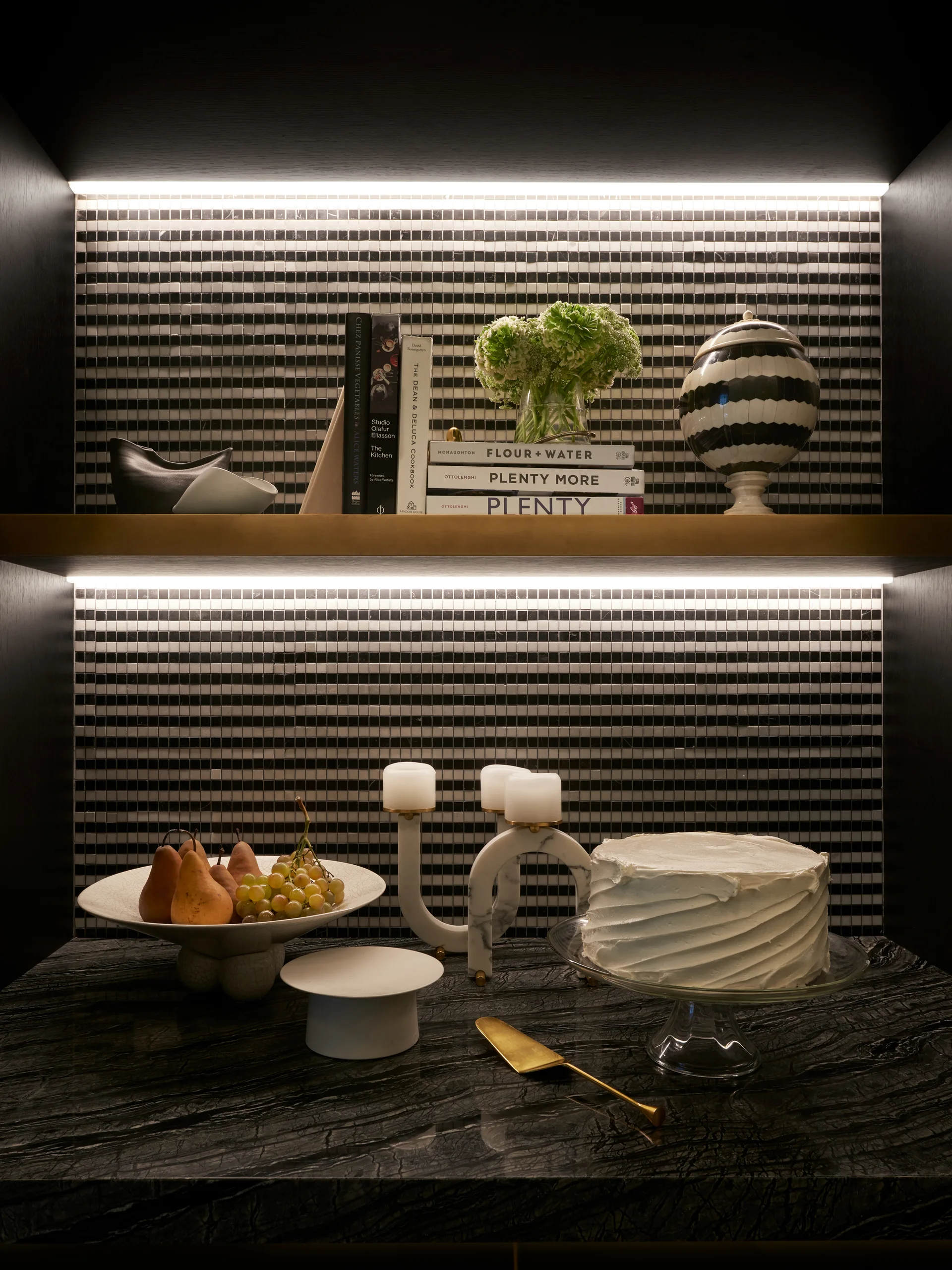 A styled shelf and countertop display featuring a black and white mosaic tile backsplash, integrated LED lighting, and cookbooks.
